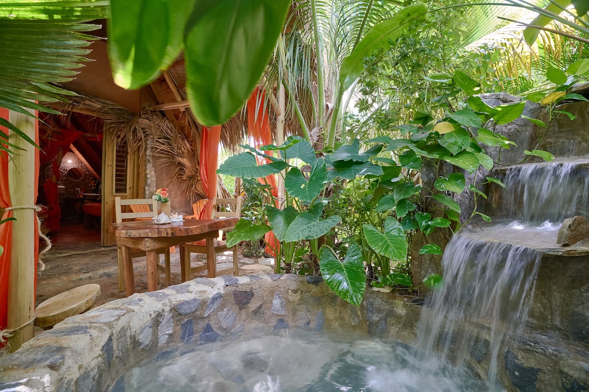 A rustic wooden table with chairs, a small waterfall, and lush greenery.