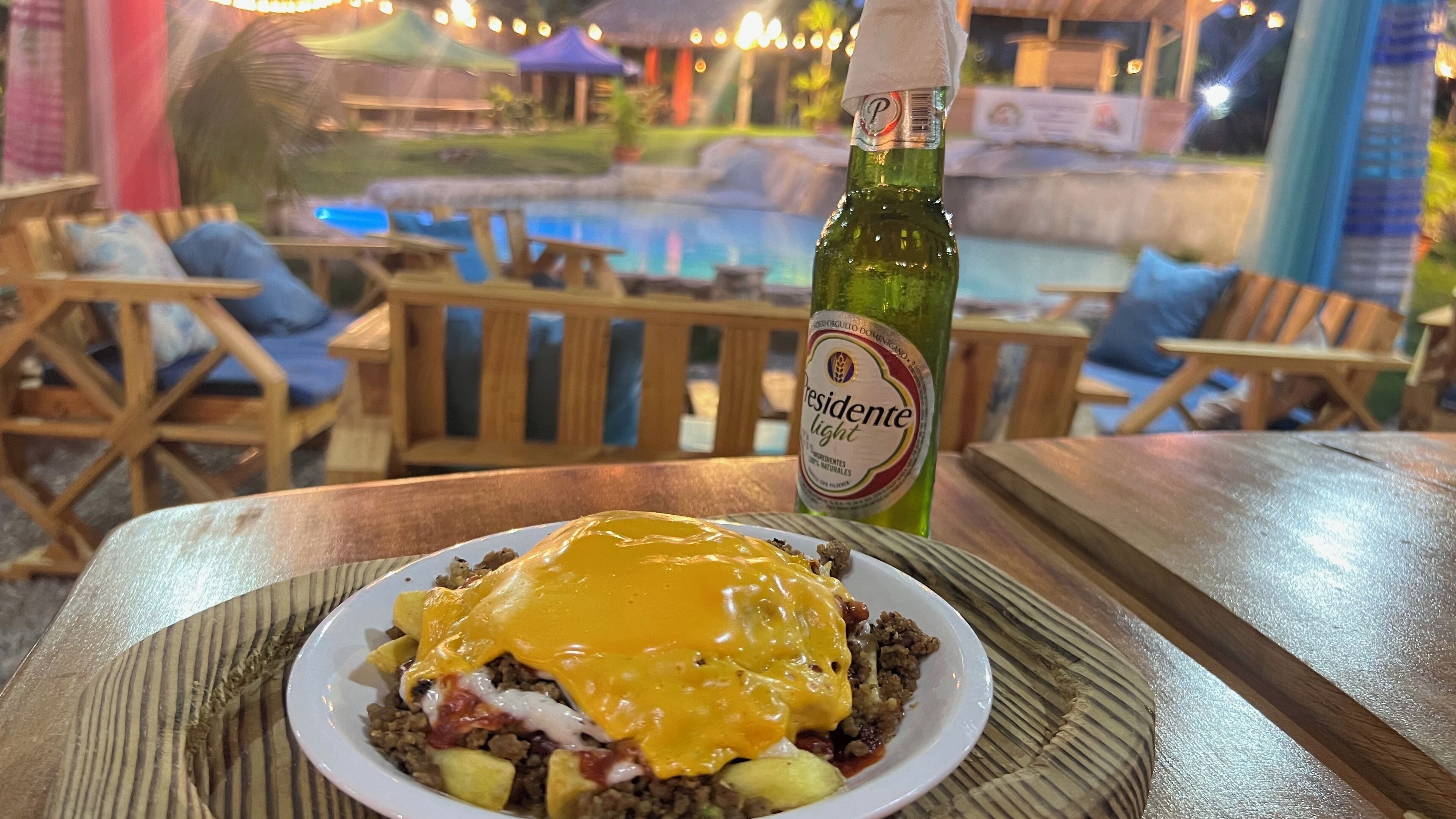 A plate of food with cheese and meat, a bottle of beer, and a poolside setting with wooden furniture.