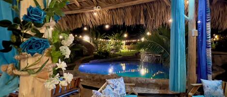 A pool area with a thatched roof, lounge chairs, and a decorative floral arrangement.