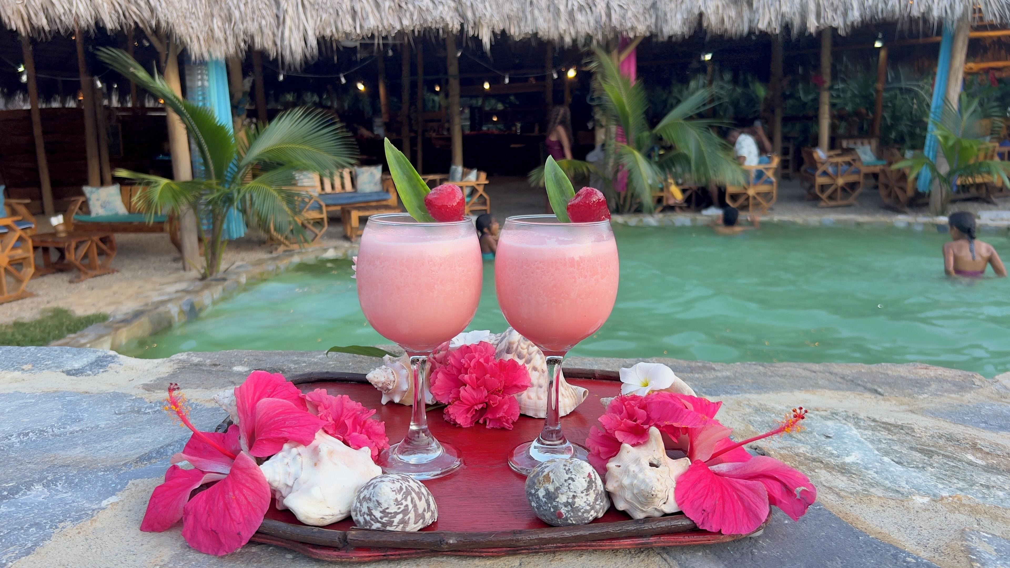 A tropical setting with a pool, lounge chairs, and a table with drinks and decorative shells.