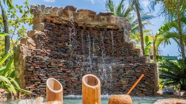 A tropical setting with a stone waterfall, a wooden tray with food and drinks, and palm trees in the background. -  CHALET TROPICAL Hotel & Pool Bar Restaurant (Las Galeras)