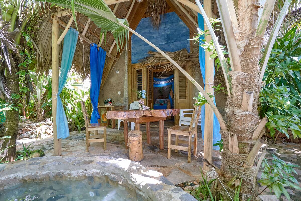 A rustic wooden structure with a thatched roof, a stone table, and wooden chairs.