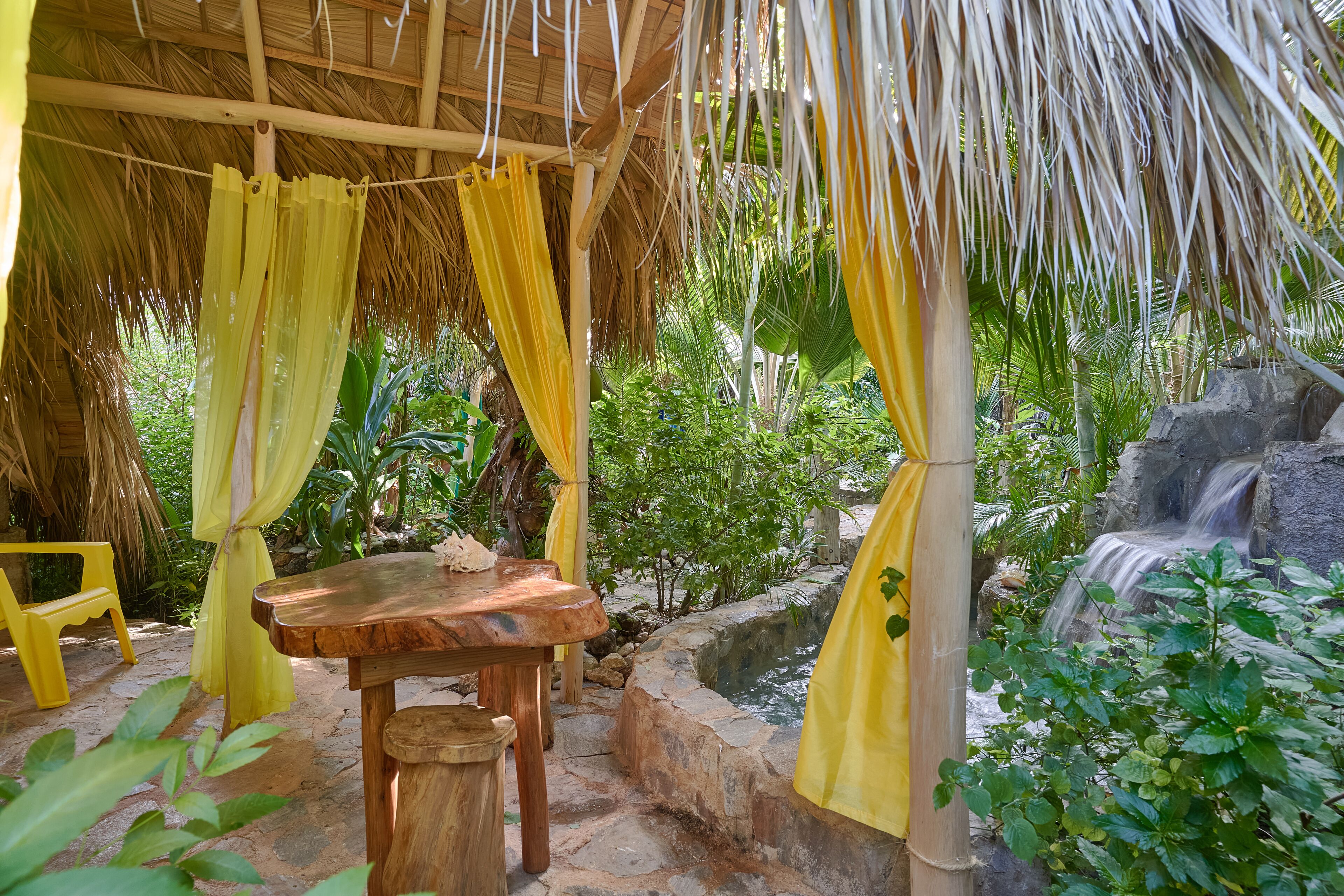 A wooden table and chair under a thatched roof with yellow curtains, surrounded by lush greenery and a small waterfall.