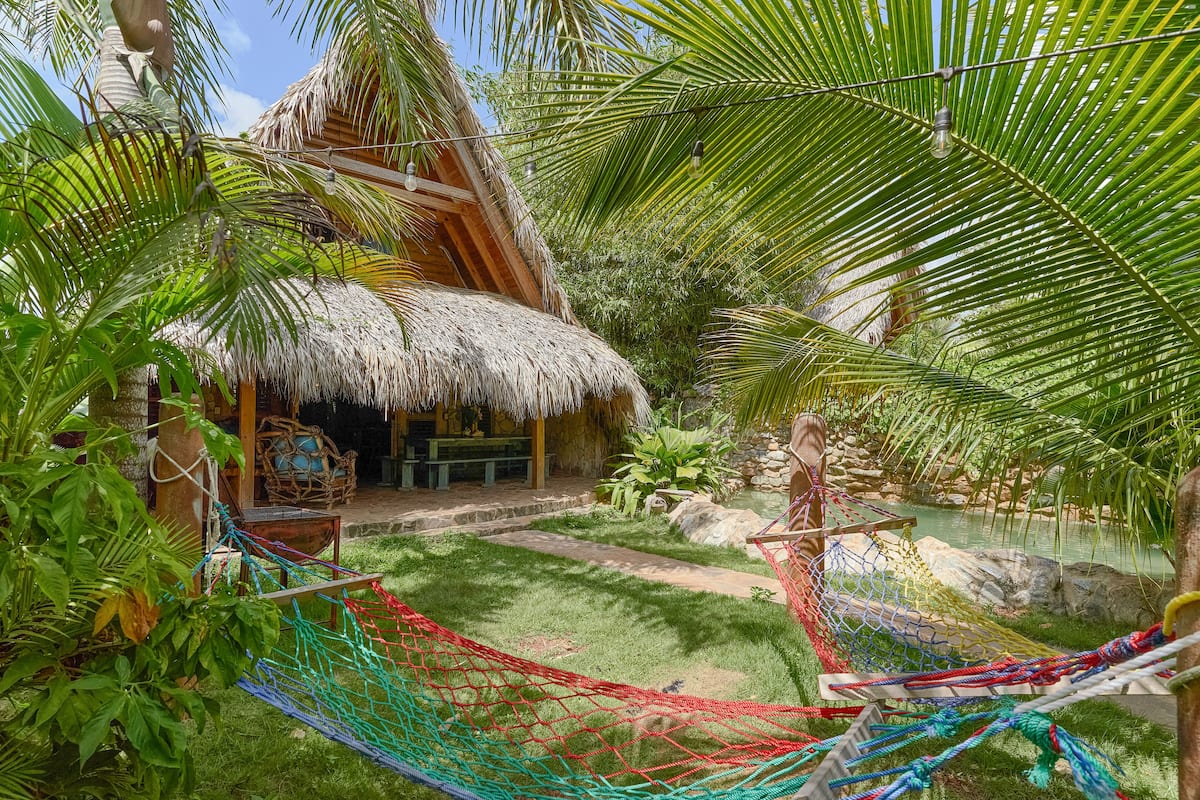 A thatched-roof hut with a dining area, surrounded by lush greenery and a hammock.