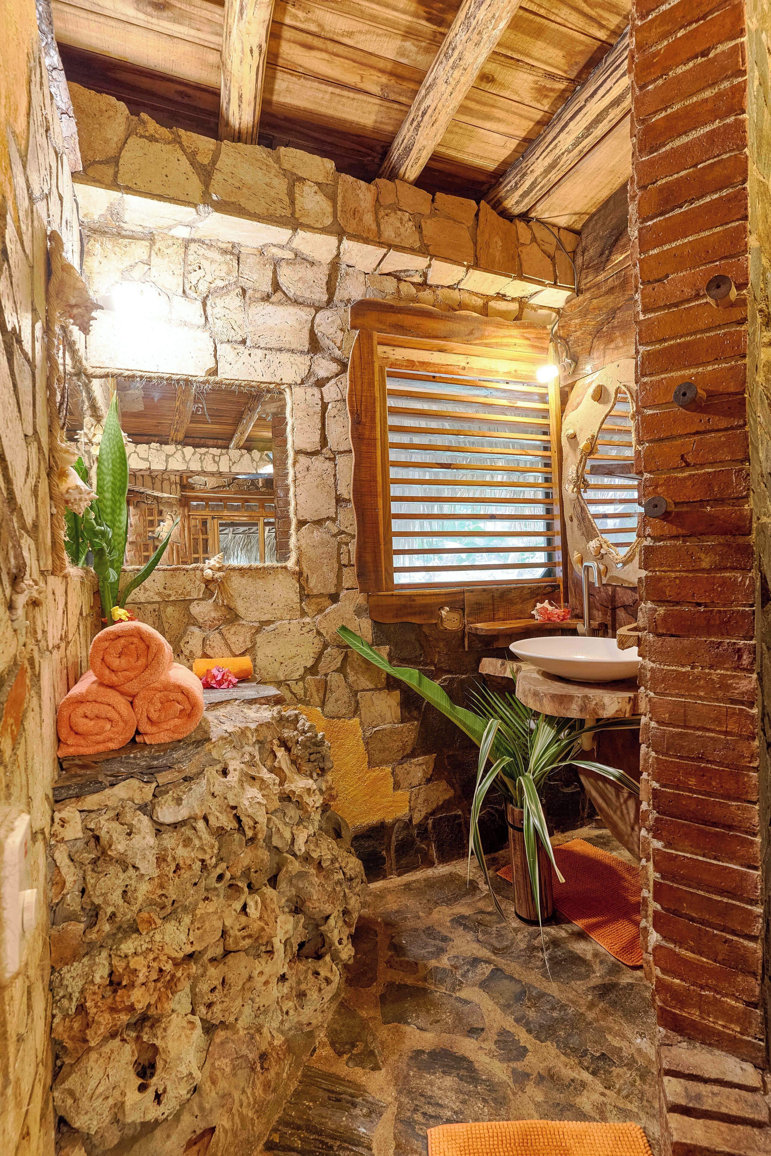 Un baño rústico con paredes de piedra, techo de madera, lavamanos de piedra y una ventana con persianas.