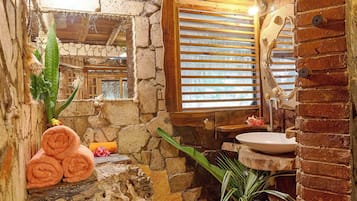 Un baño rústico con paredes de piedra, techo de madera, lavamanos de piedra y una ventana con persianas.