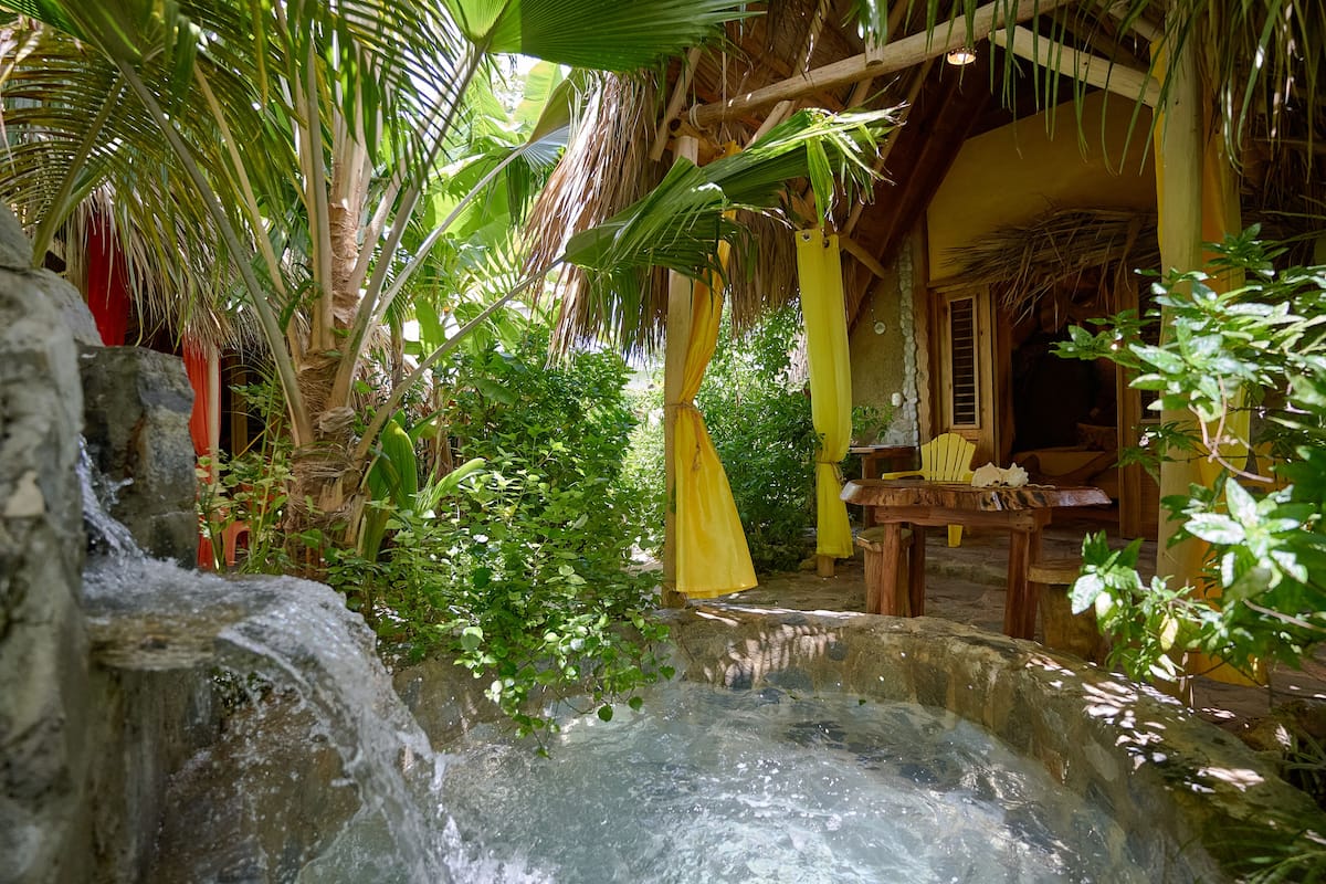 A tropical setting with a stone water feature, wooden furniture, and a thatched-roof structure.