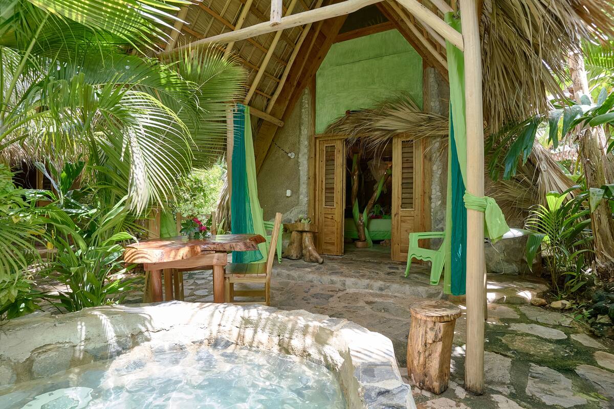 A rustic wooden structure with a thatched roof, stone flooring, and a small water feature.