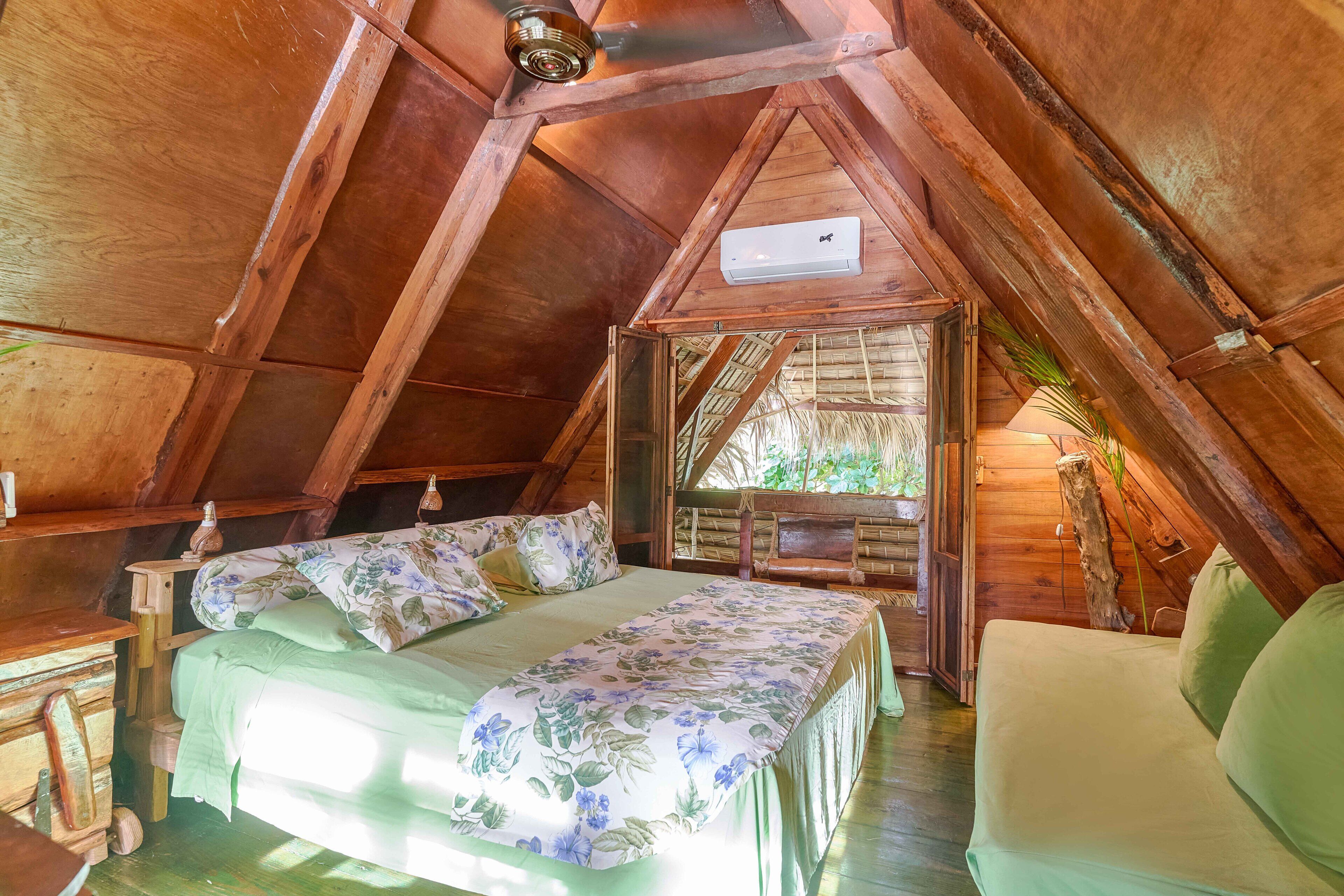 A cozy attic bedroom with a wooden ceiling, a bed with floral bedding, a chair, and a small table.