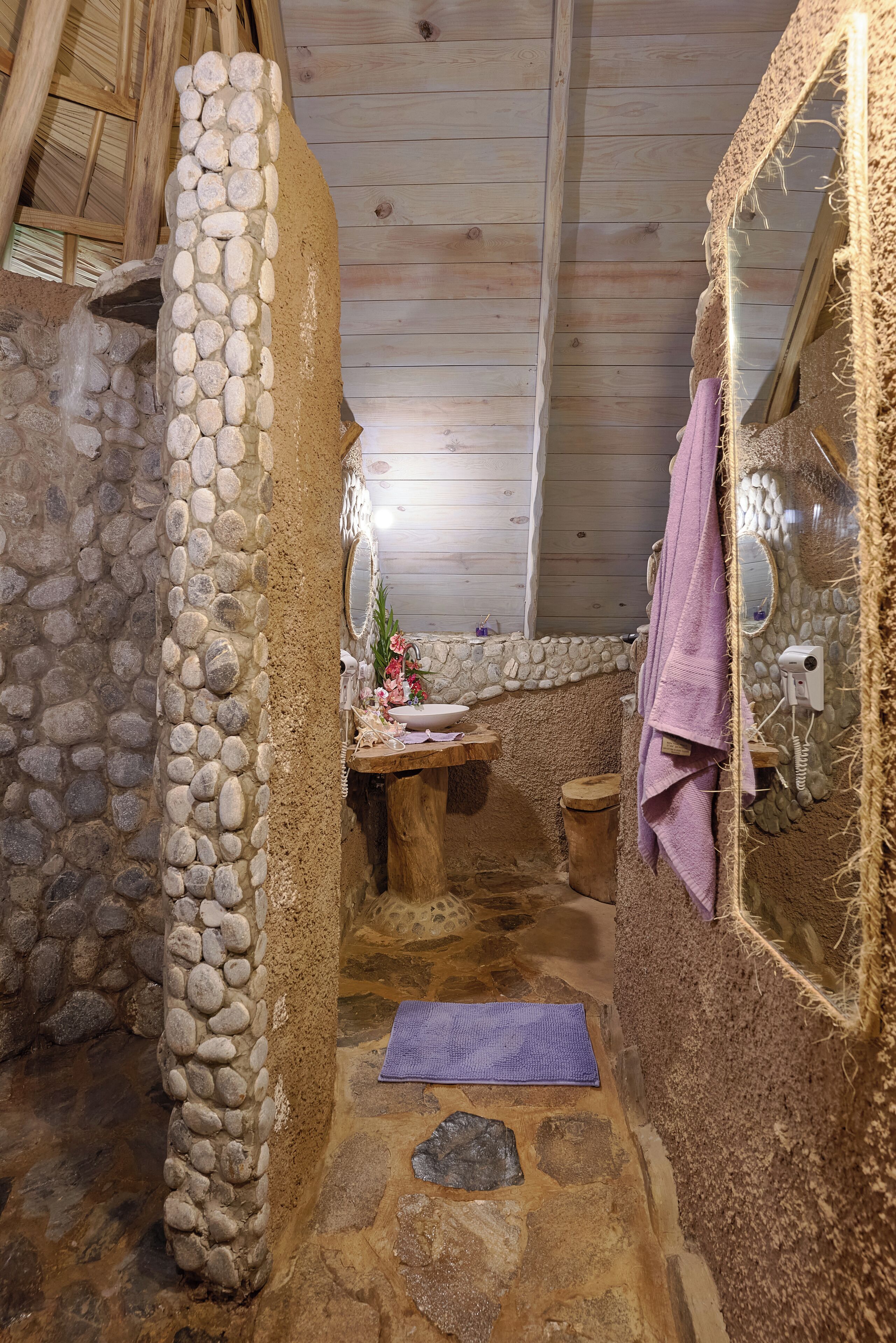 A rustic bathroom with stone walls, a wooden bench, a purple mat, and a mirror with a textured frame.