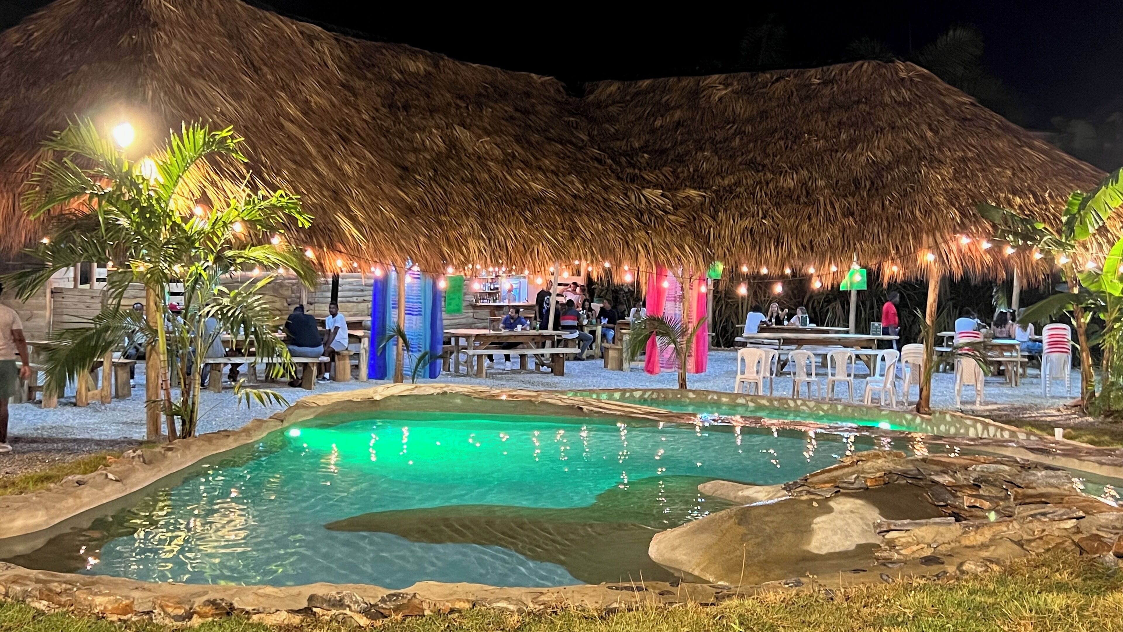 An outdoor pool area with a thatched-roof structure, surrounded by tables and chairs, and a small pool with rocks and pebbles.