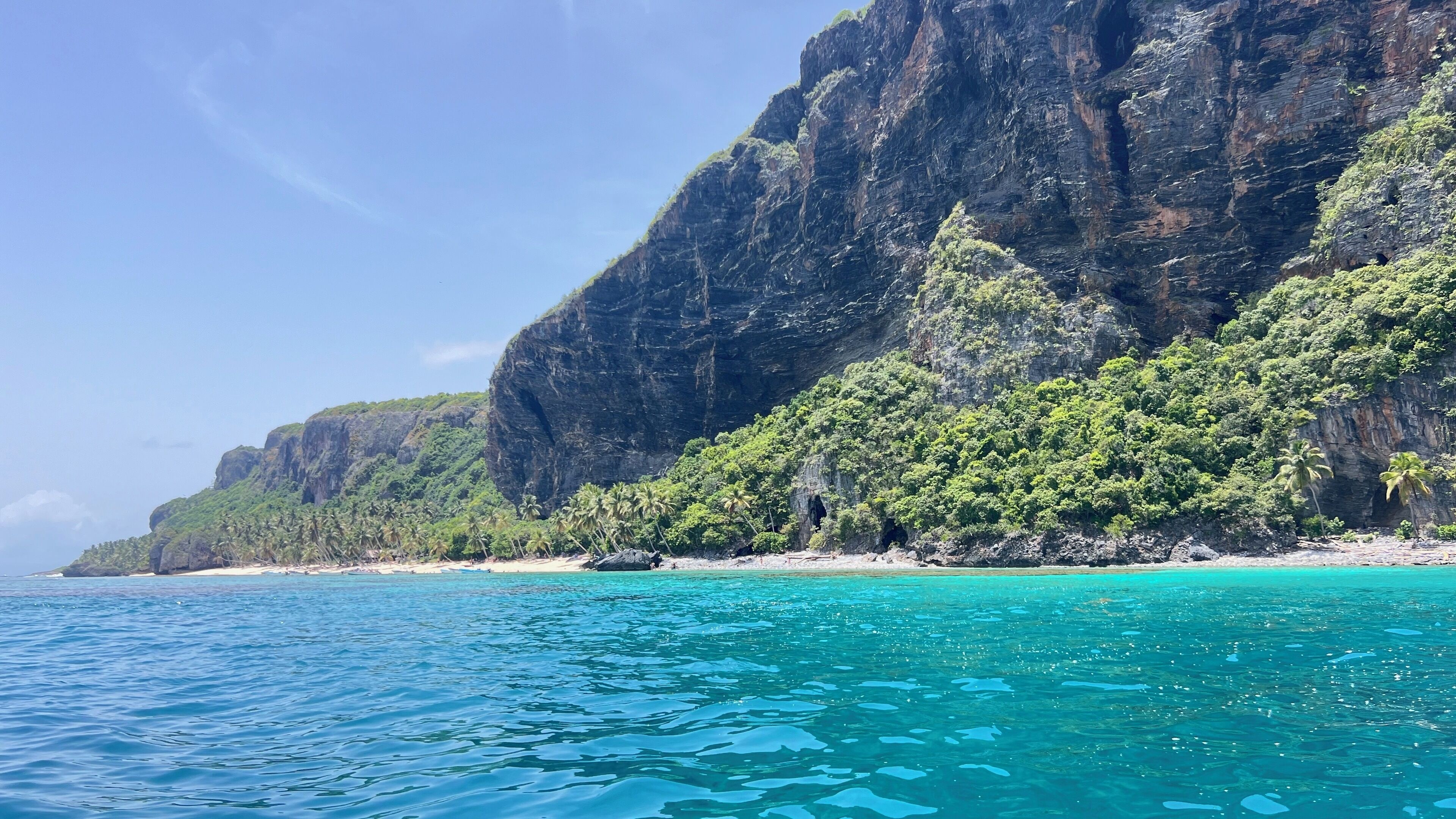 Un paysage côtier caractérisé par une eau turquoise cristalline, une plage de sable et une falaise abrupte couverte d’une végétation luxuriante.