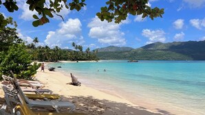 A beach with clear turquoise water, palm trees, and a mountain in the background. -  CHALET TROPICAL Hotel & Pool Bar Restaurant (Las Galeras)