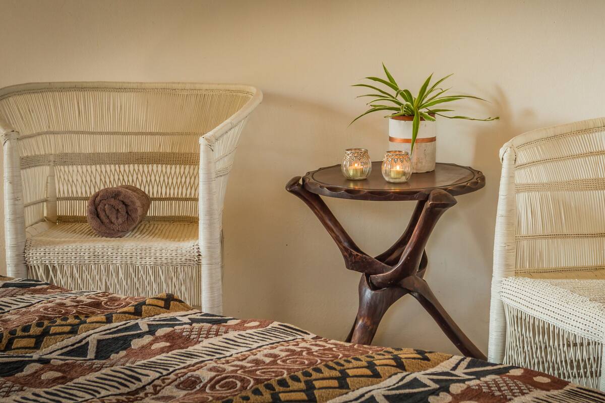 Une pièce comprenant deux fauteuils en osier, une petite table ronde ornée d’une plante et de bougies, et un couvre-lit à motifs.