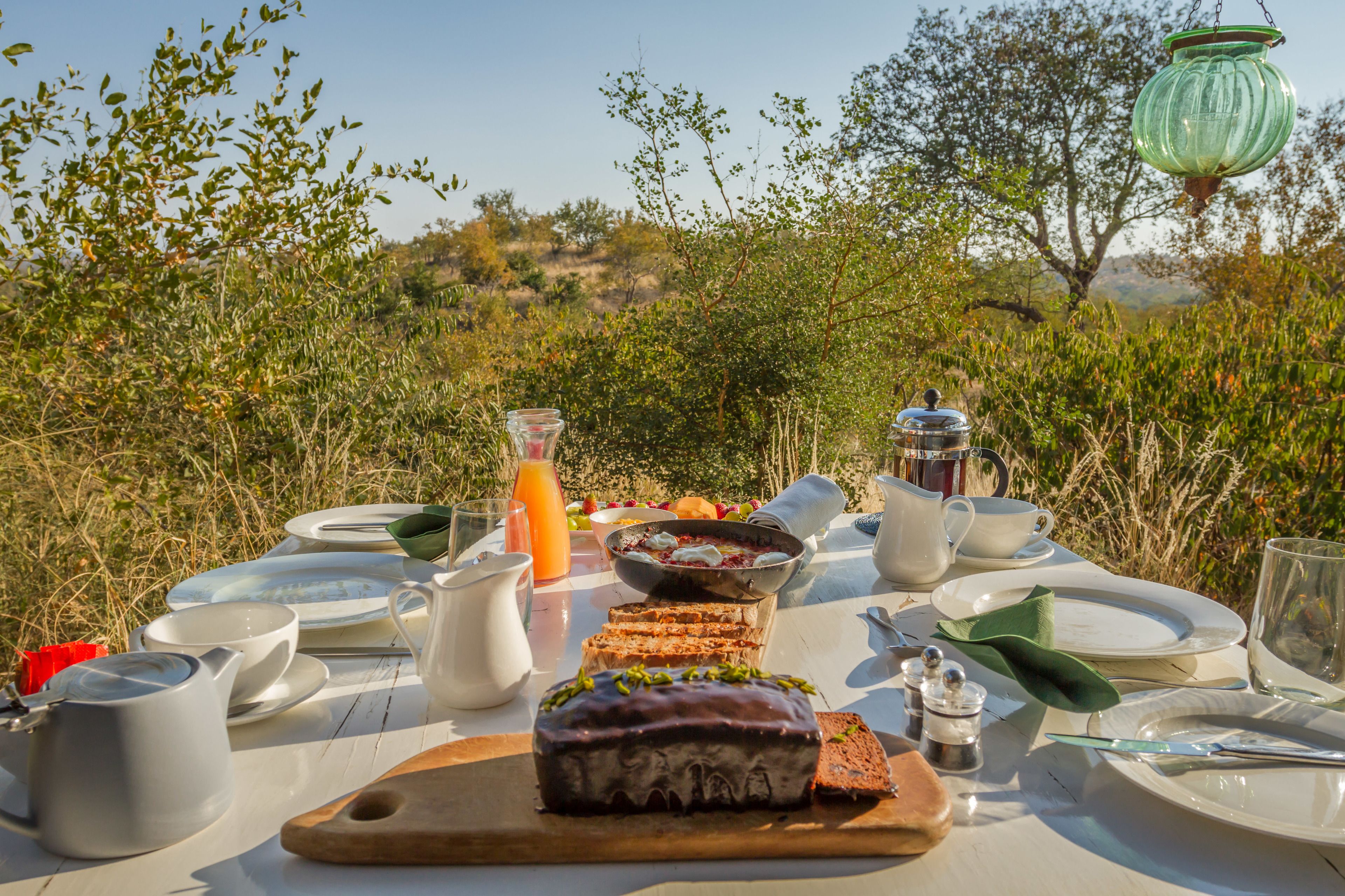 Une table dressée à l’extérieur avec des aliments pour le petit-déjeuner, notamment une miche de pain, une carafe de jus d’orange et divers plats.