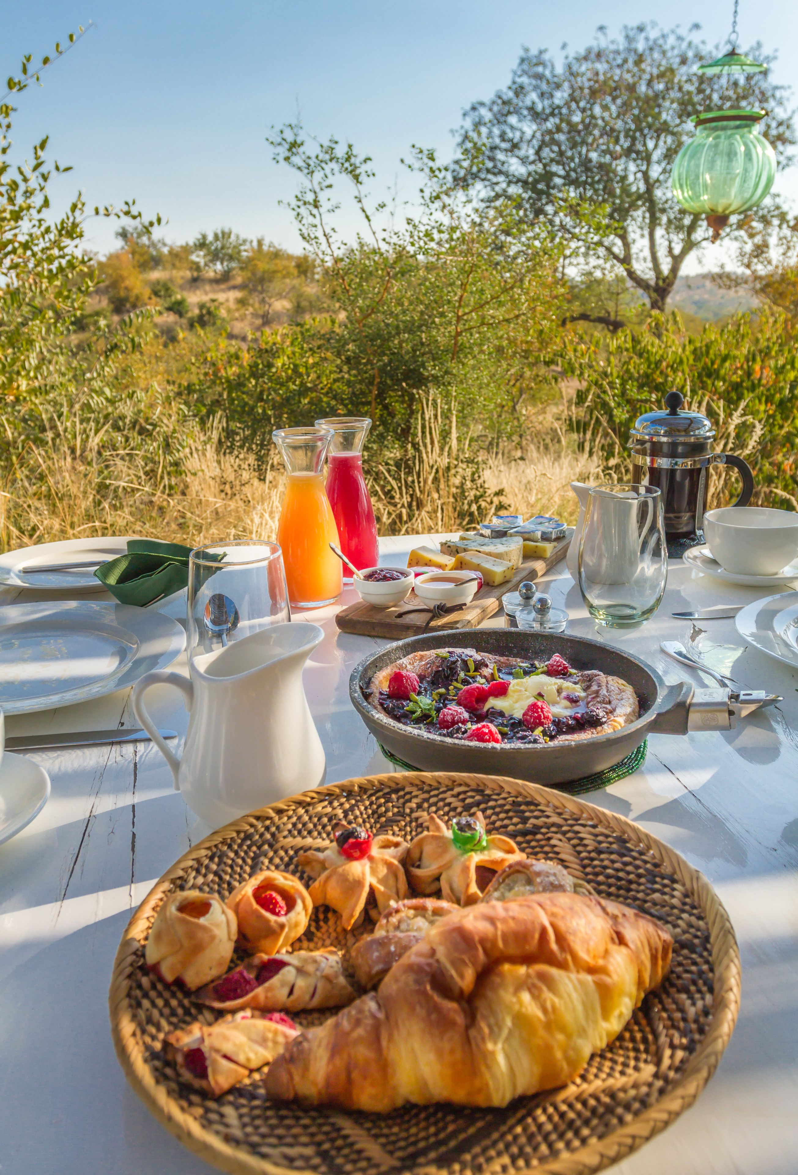 Una mesa de desayuno al aire libre con diversos alimentos, incluyendo pasteles y un bol de frutas, además de una cafetera.