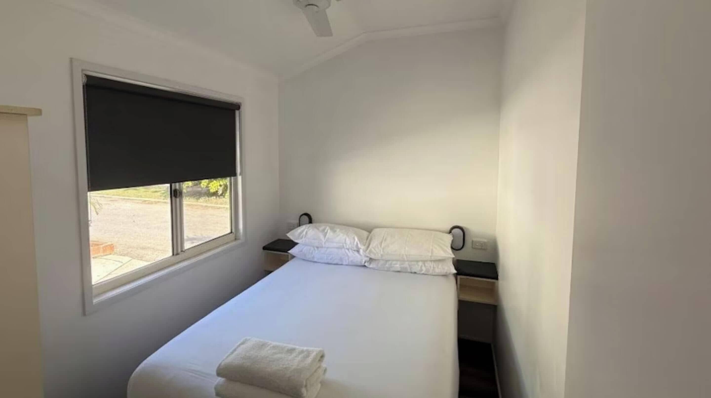 A small, single-bed room with a window, a bedside table, and a ceiling fan.
