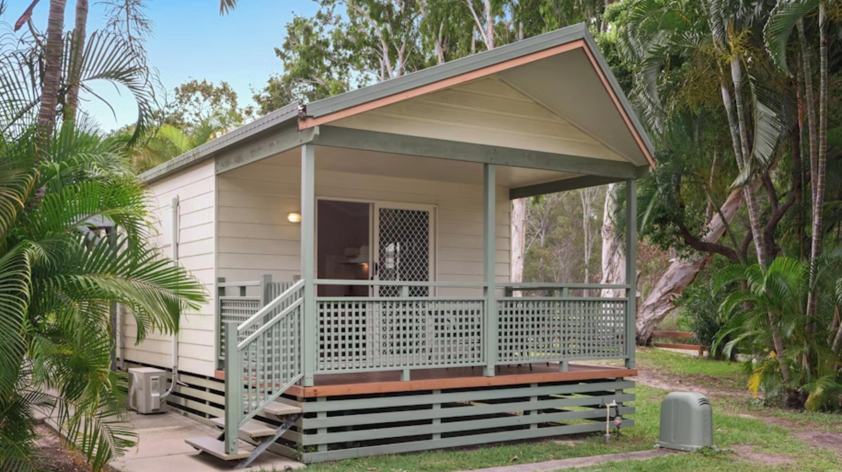 A small, single-story cabin with a porch and a covered area, surrounded by lush greenery.