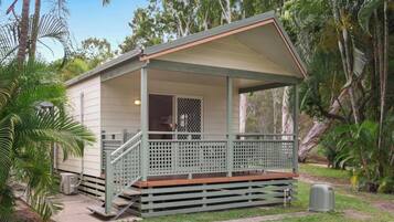 A small, single-story cabin with a porch and a covered area, surrounded by lush greenery.