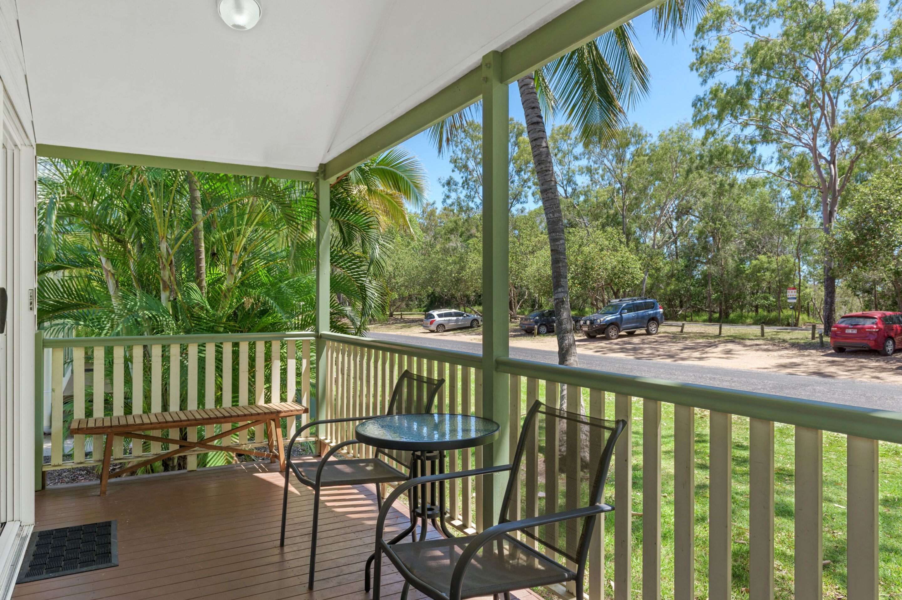A covered outdoor area with a wooden table and chairs, surrounded by a green railing and palm trees.