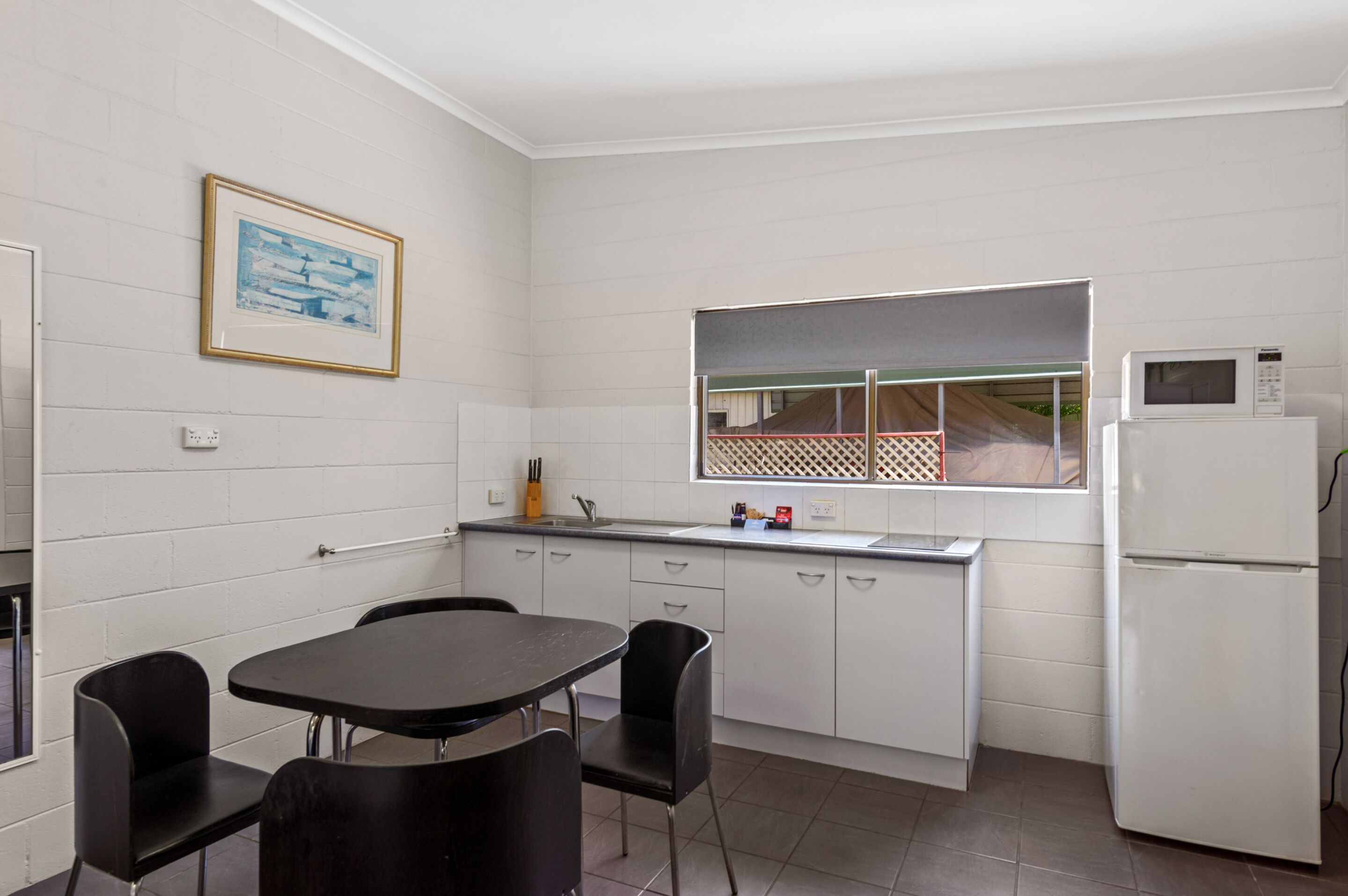A compact kitchen with white cabinets, a black dining table, and chairs. There is a window with a view of a fence and a microwave on the counter.