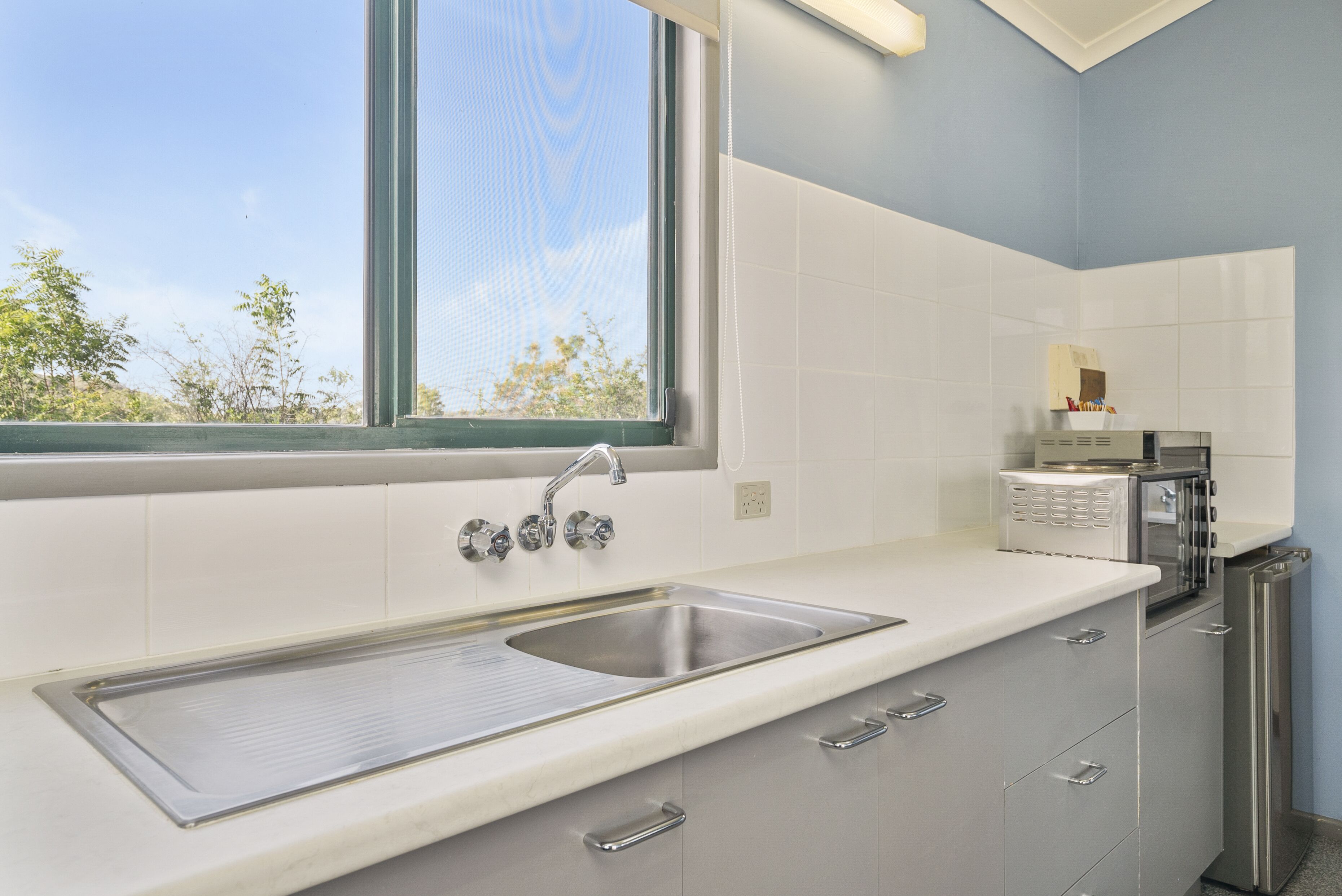 A modern kitchen with a stainless steel sink, white cabinets, a built-in oven, and a window with a view of trees.