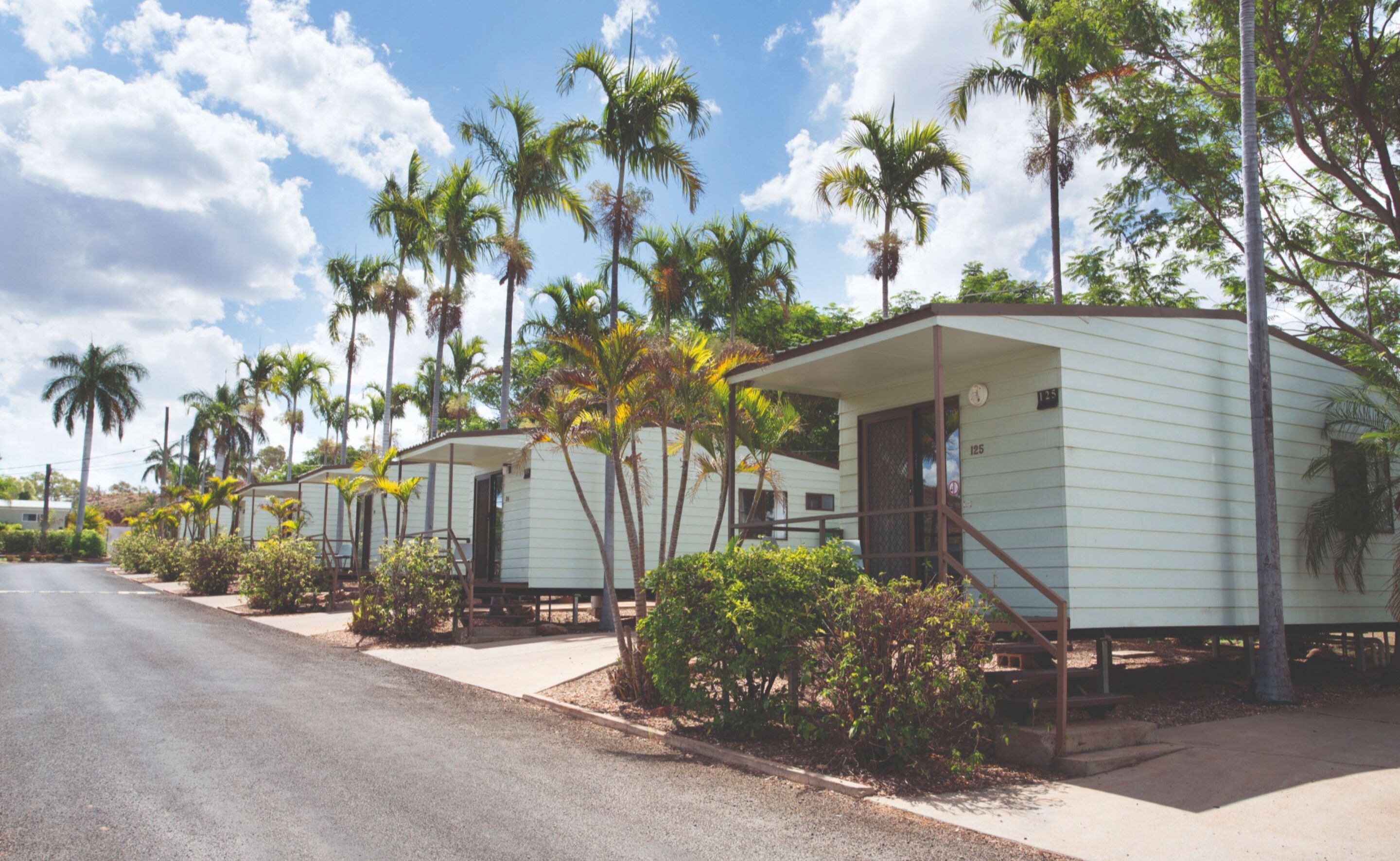 A row of mobile homes with a paved road in front, surrounded by palm trees and greenery.