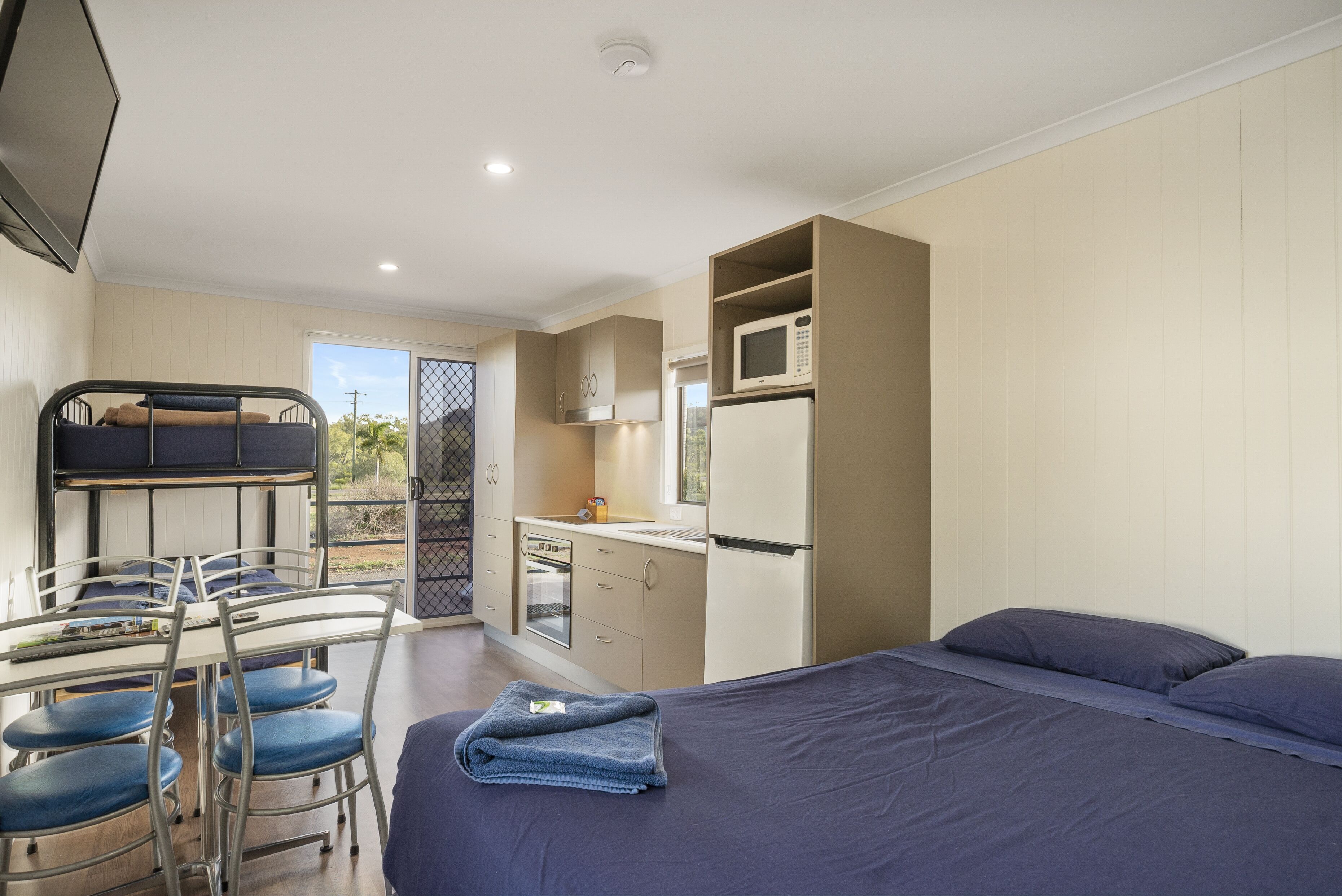 A compact room with a bed, a dining table with chairs, a kitchenette, and a TV.