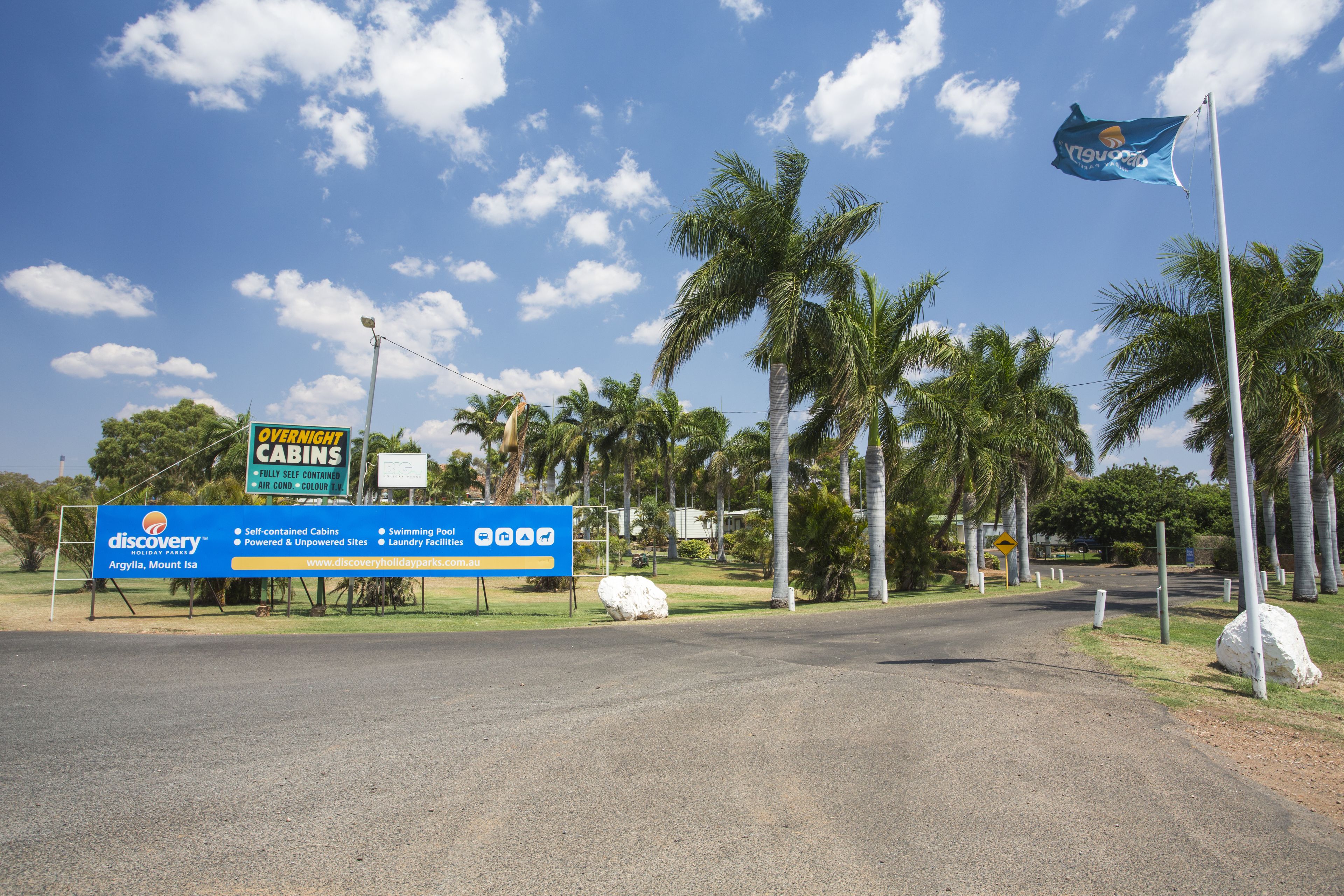 Uma estrada leva a um parque de férias, com um letreiro anunciando cabanas completas, ar condicionado e piscina.