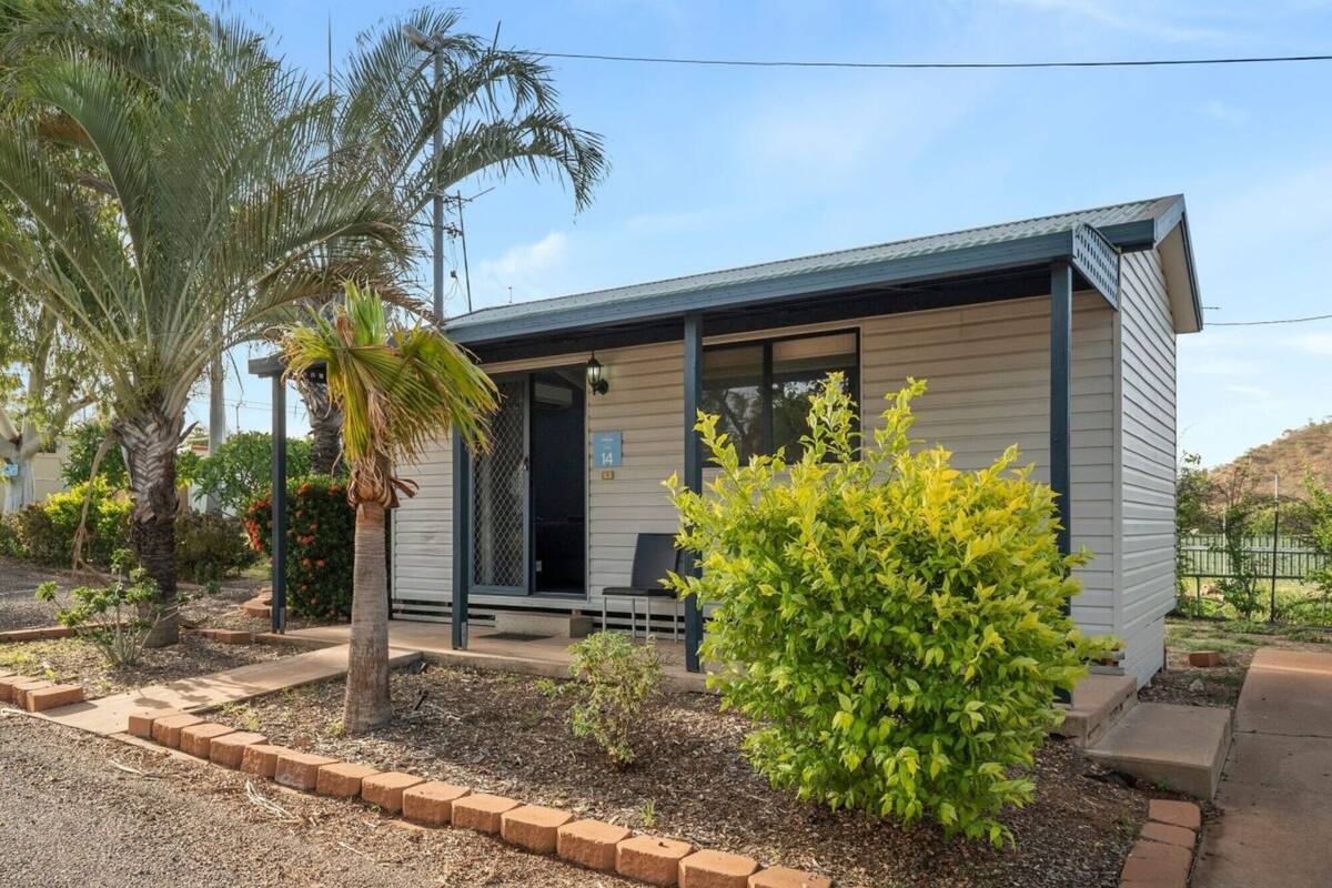 A small, single-story building with a covered porch, a number '14' sign, and a well-maintained garden with palm trees and shrubs.