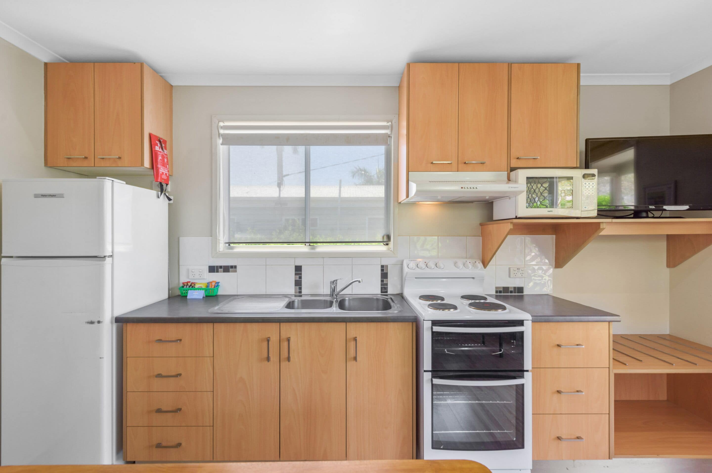 A modern kitchen with wooden cabinets, a built-in microwave, a refrigerator, and a window with blinds.