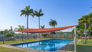 Una piscina rectangular con una vela roja de sombra sobre ella, rodeada por una zona vallada con palmeras y un césped.