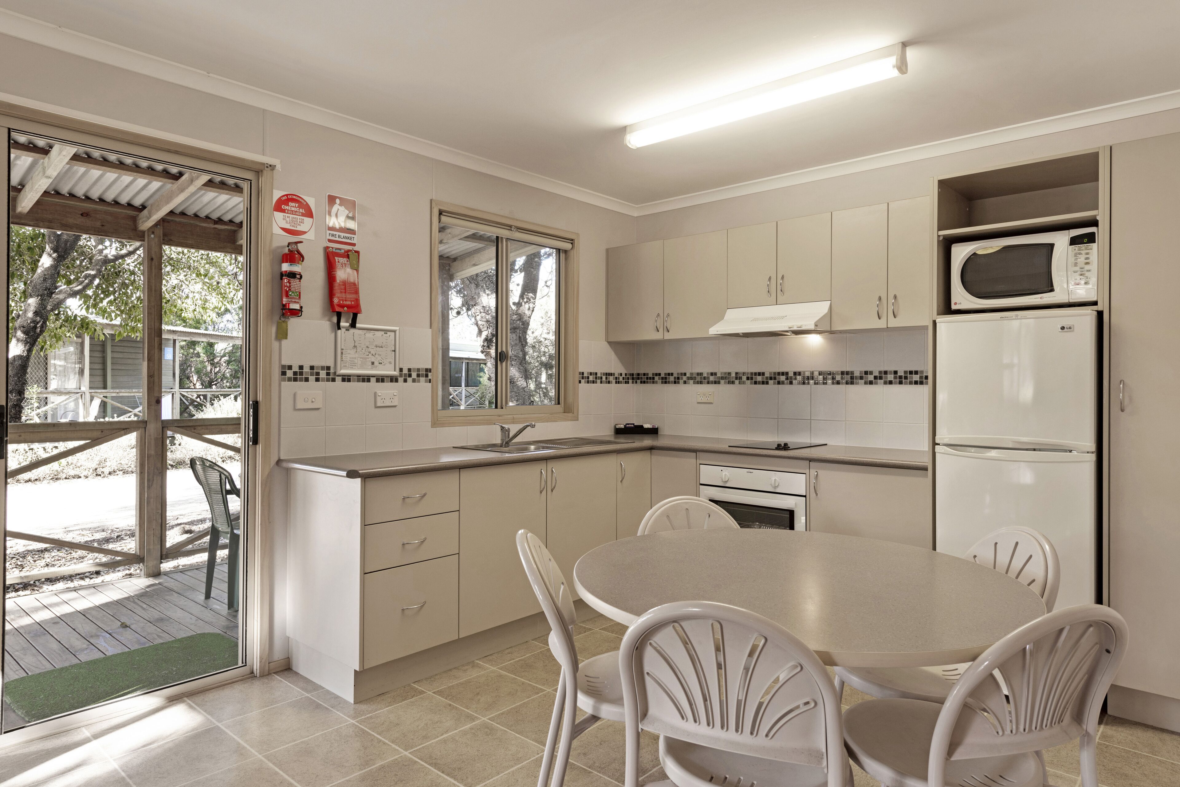 A modern kitchen with white cabinets, a built-in microwave, and a refrigerator. There is a dining table with four chairs and a window providing natural light.