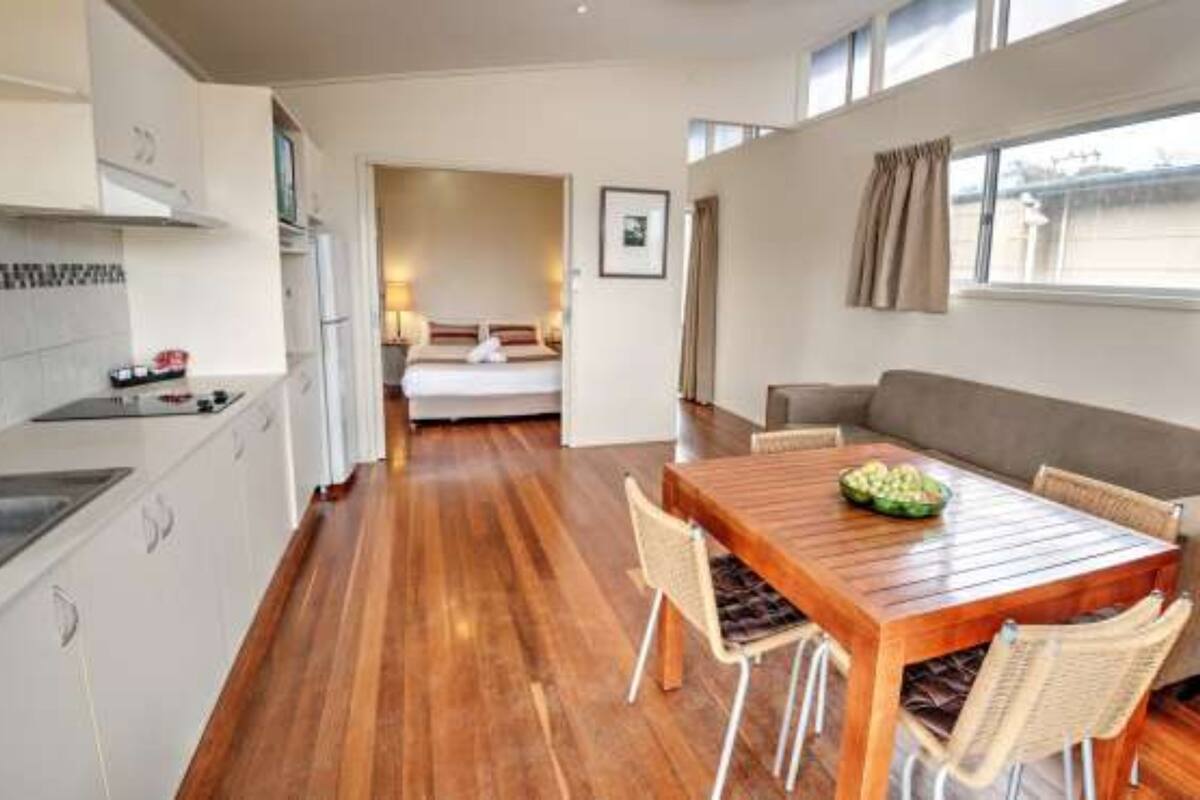 A compact hotel room with a kitchenette, a dining area with a wooden table and chairs, a bed with a view, and a window with curtains.