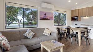 Una sala moderna con sofá, una mesa de centro y un comedor con sillas y una mesa. Hay ventanas amplias que ofrecen vistas a árboles y un cuadro de un faro.