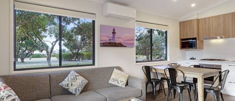 A modern living room with a sofa, coffee table, and dining area with chairs and a table. There are large windows with a view of trees and a painting of a lighthouse.