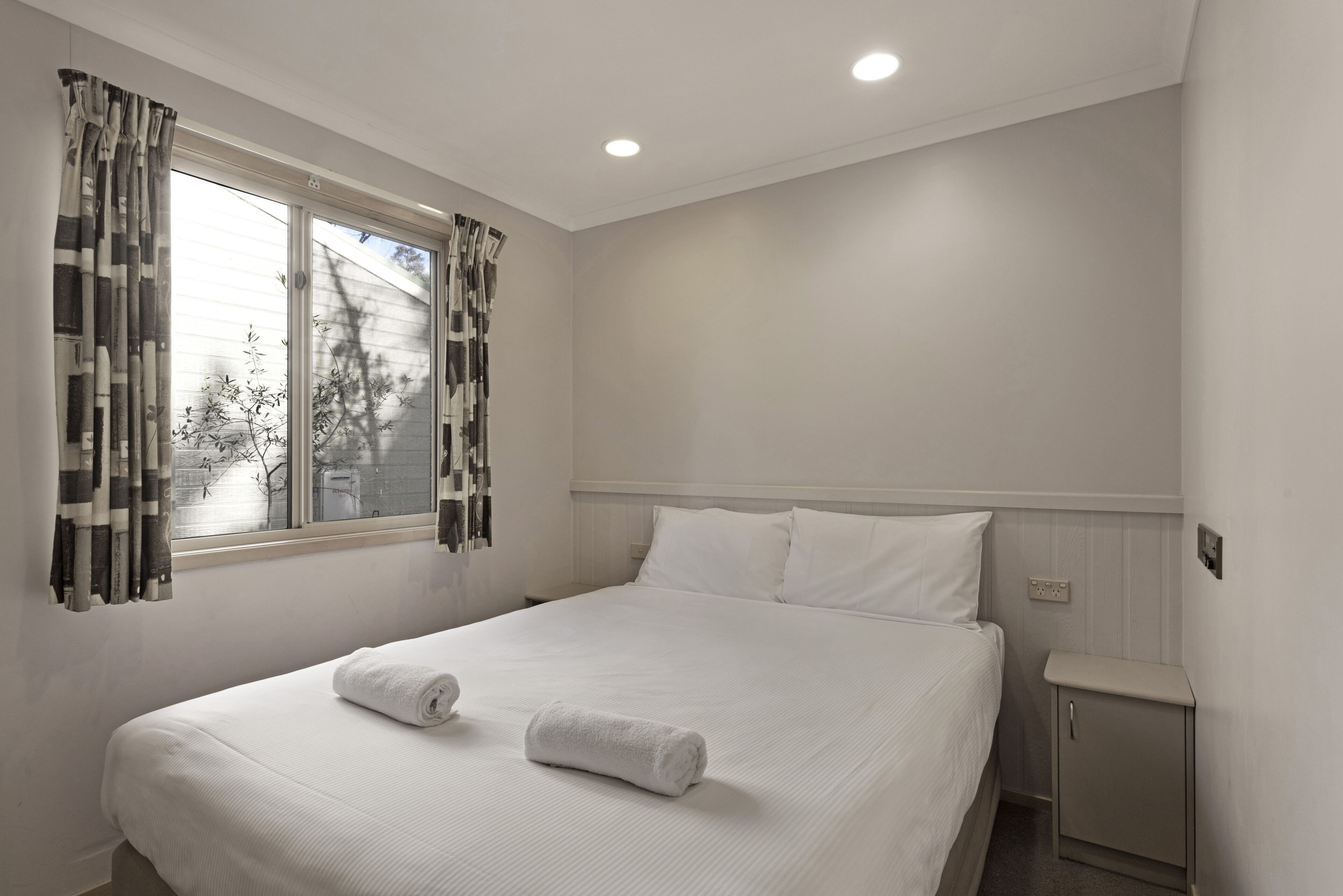 Una cama bien hecha con sábanas blancas y dos toallas enrolladas, una mesita de noche con lámpara, y una ventana con cortinas.