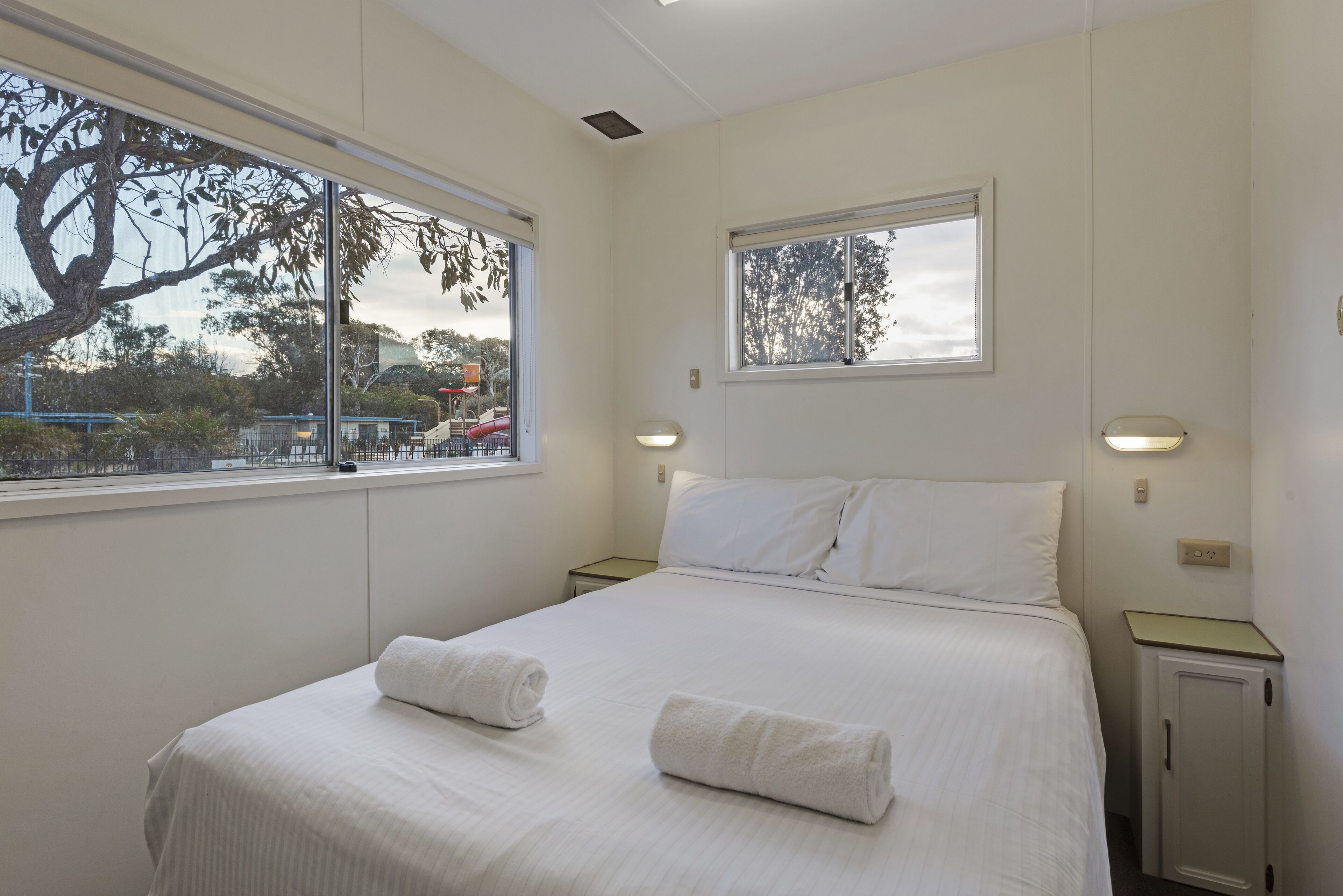 Una habitación de hotel con una cama grande, dos mesitas de noche, una ventana con vistas a árboles y un parque infantil, y un mueble pequeño.