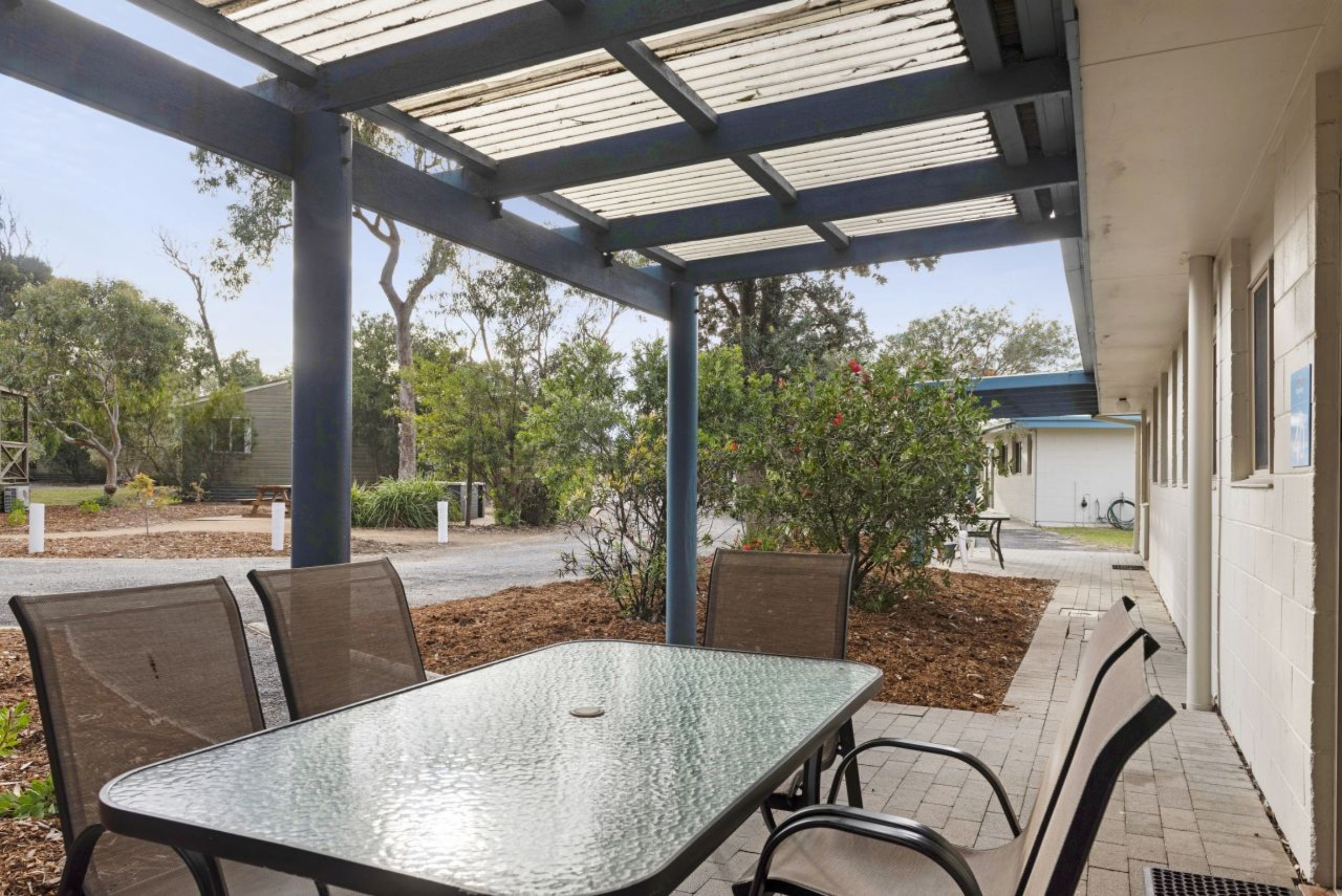 A covered outdoor seating area with a table and chairs, surrounded by trees and a building.