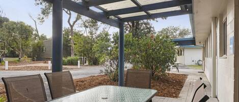 A covered outdoor seating area with a table and chairs, surrounded by trees and a building.
