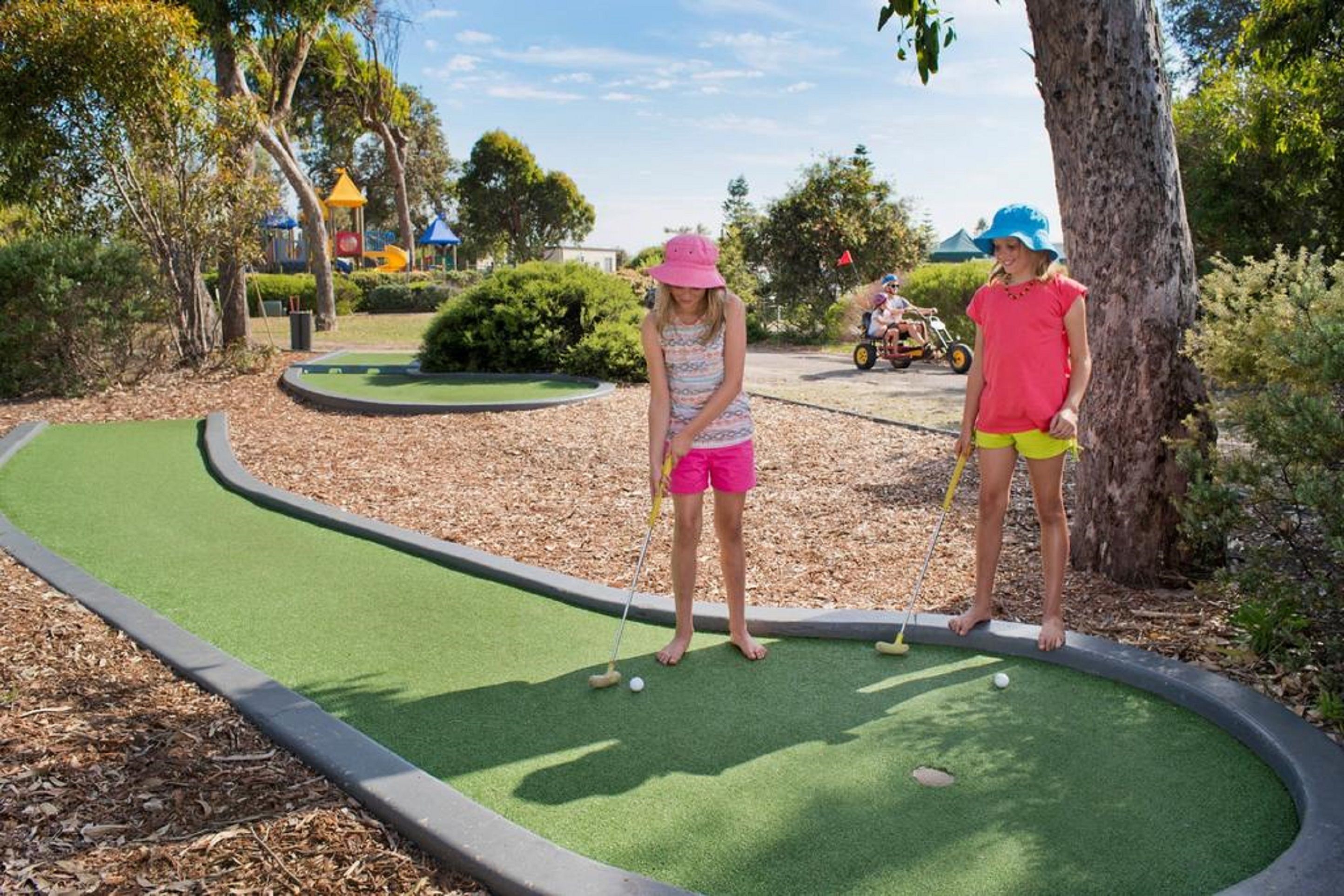 A miniature golf course with a green putting surface, surrounded by trees and a playground in the background.