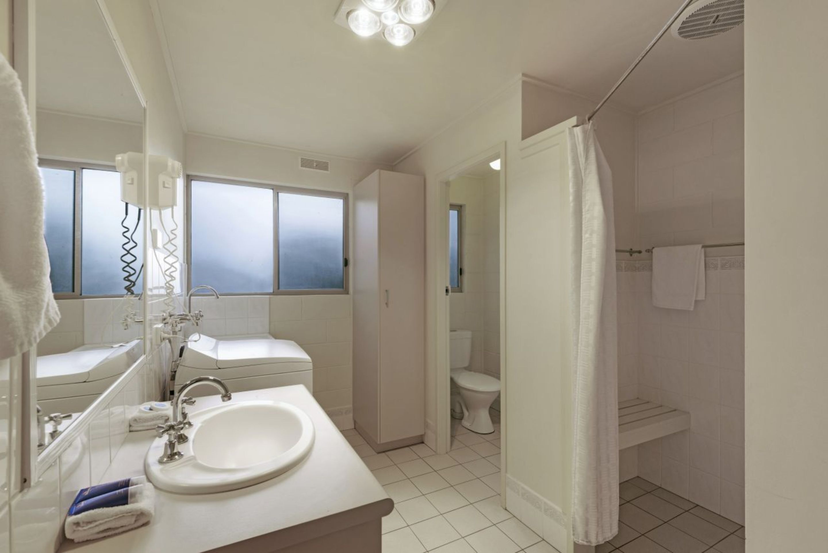 A modern bathroom with a white sink, mirror, and toilet. There are windows with frosted glass, a shower curtain, and a towel rack.