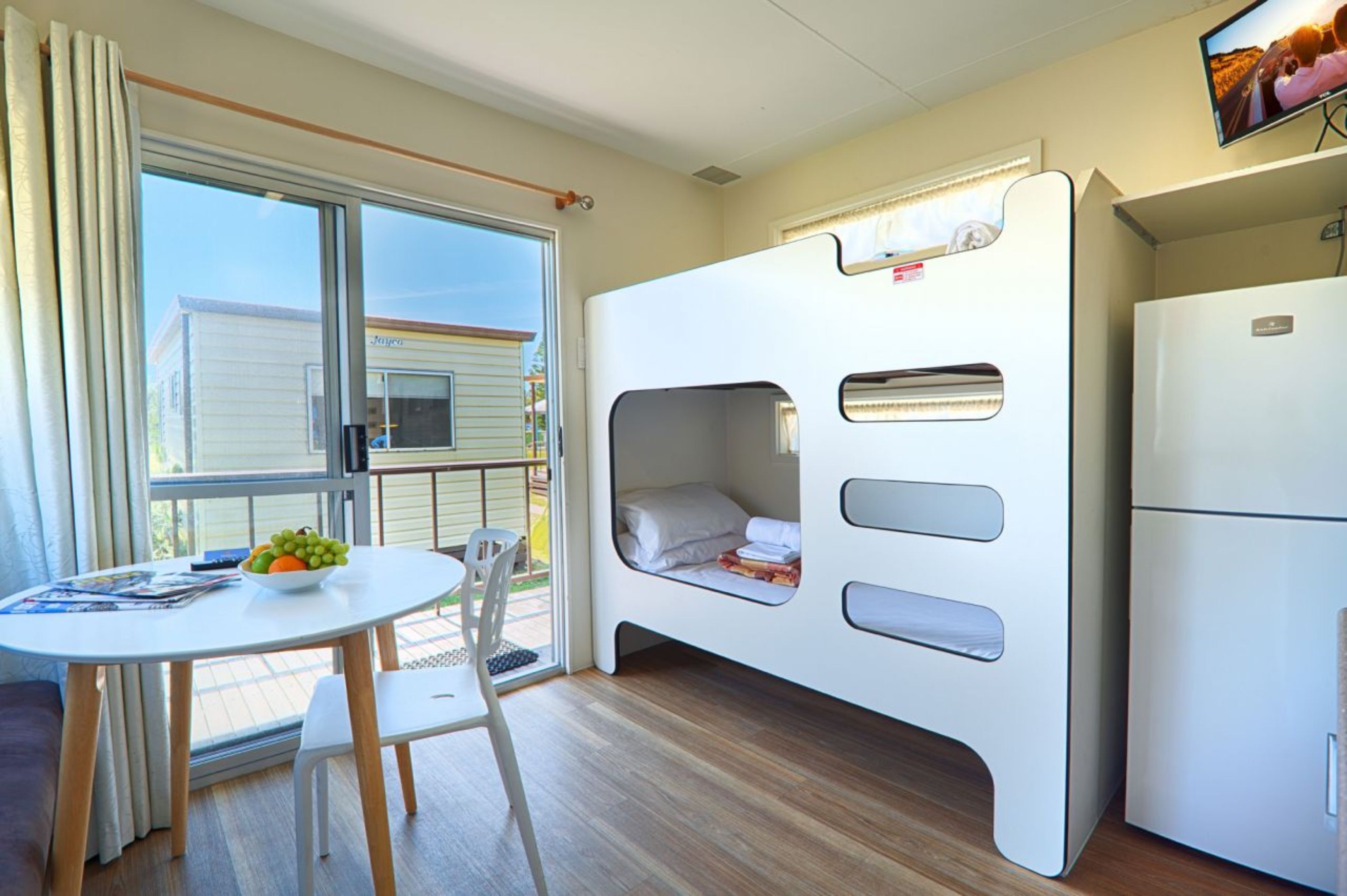 A compact room with a bunk bed, a small table with a bowl of fruit, a white refrigerator, and a TV mounted on the wall.