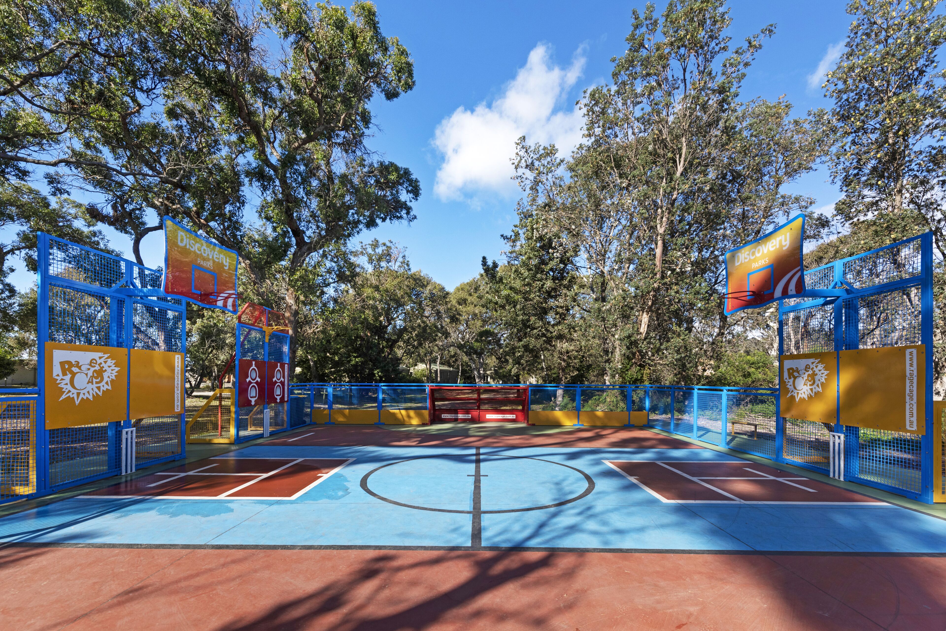 Una cancha de baloncesto al aire libre con piso azul y rojo, rodeada de vallas azules y amarillas, y árboles al fondo.