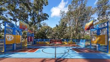 Una cancha de baloncesto al aire libre con piso azul y rojo, rodeada de vallas azules y amarillas, y árboles al fondo.