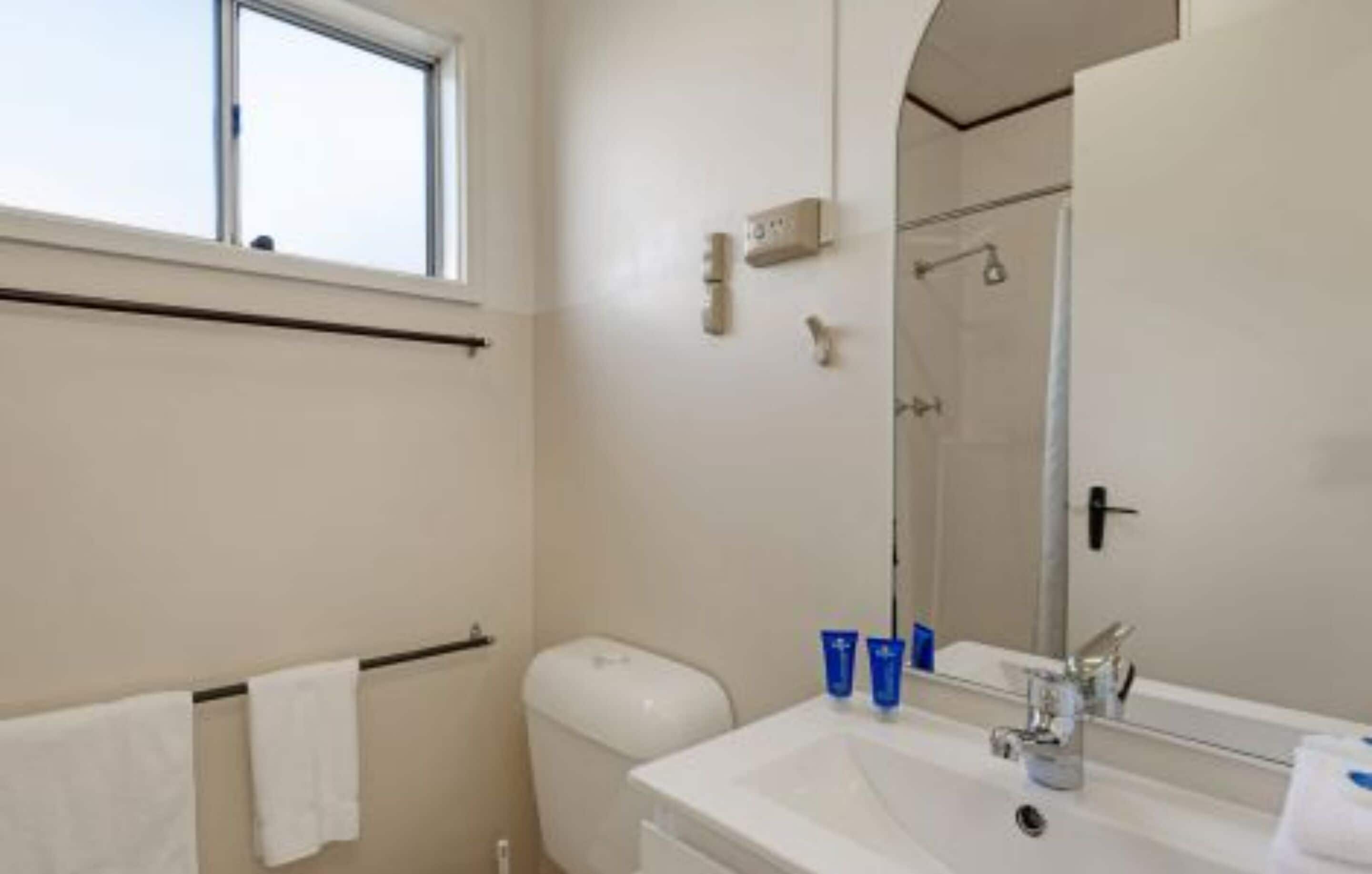 A bathroom with a toilet, sink, and shower. There is a window above the toilet and a towel rack with towels.
