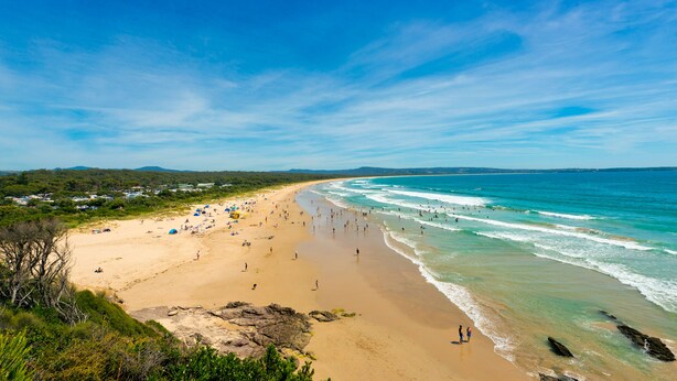 Una playa con gente y tiendas de campaña, cielos despejados y vista al océano.