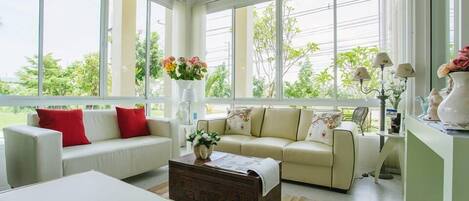 A spacious living room with a large window, a white sofa with red cushions, a wooden coffee table, and a beige sofa with floral cushions.