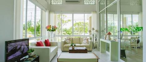 A modern living room with a sofa, coffee table, and TV. Large windows provide natural light and views of greenery outside.