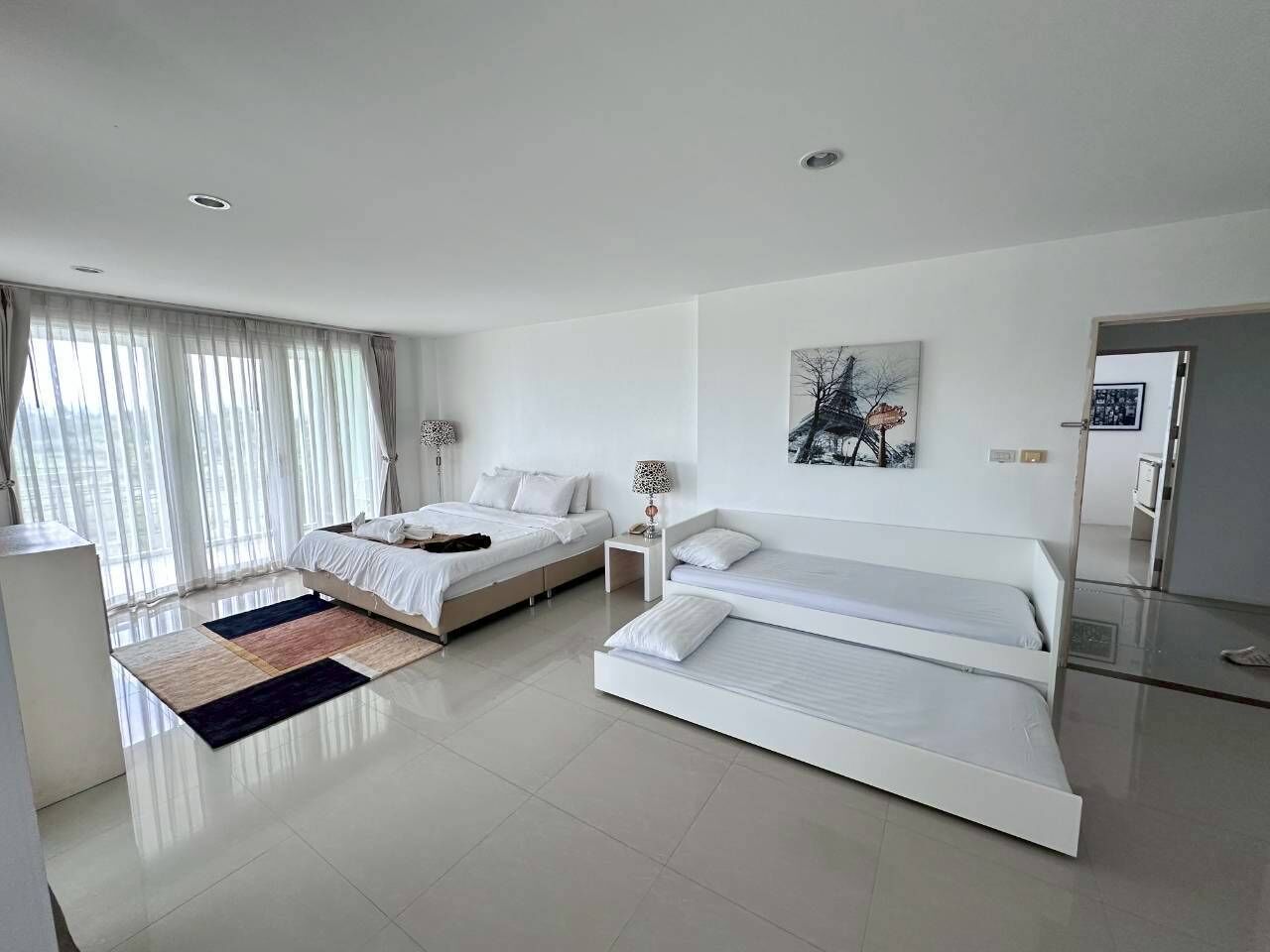 A modern bedroom with two beds, a large window with curtains, a painting on the wall, and a bedside table with a lamp.